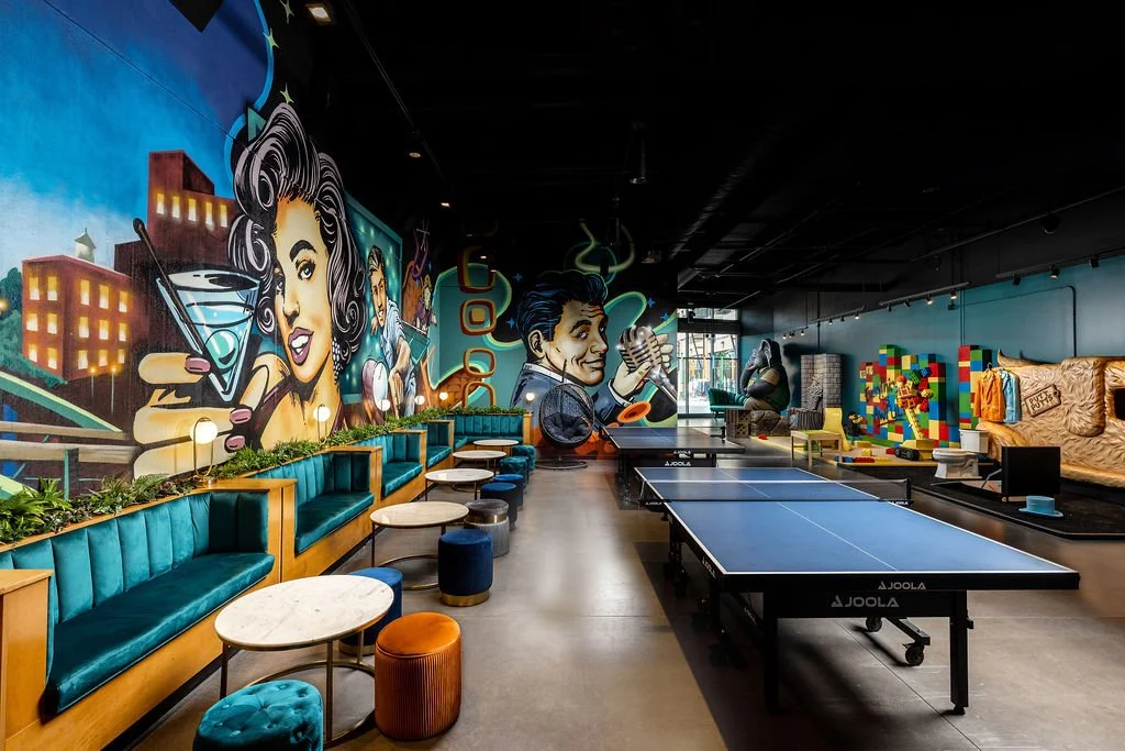 Interior of a recreational space with a colorful mural on the wall, featuring a woman painting her nails and a man drinking from a cup, ping pong tables, lounge seating, and playful decor.