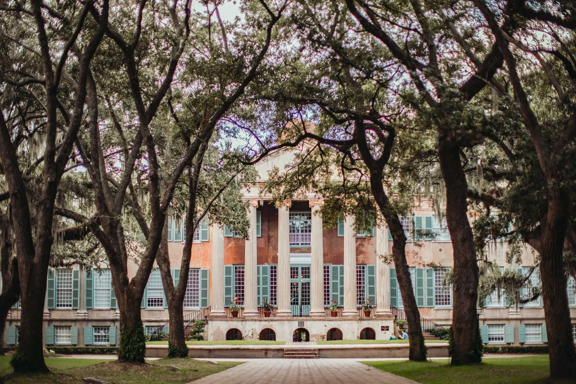 A large building with tall white columns and numerous windows, partially obscured by tall, leafy trees in the foreground.