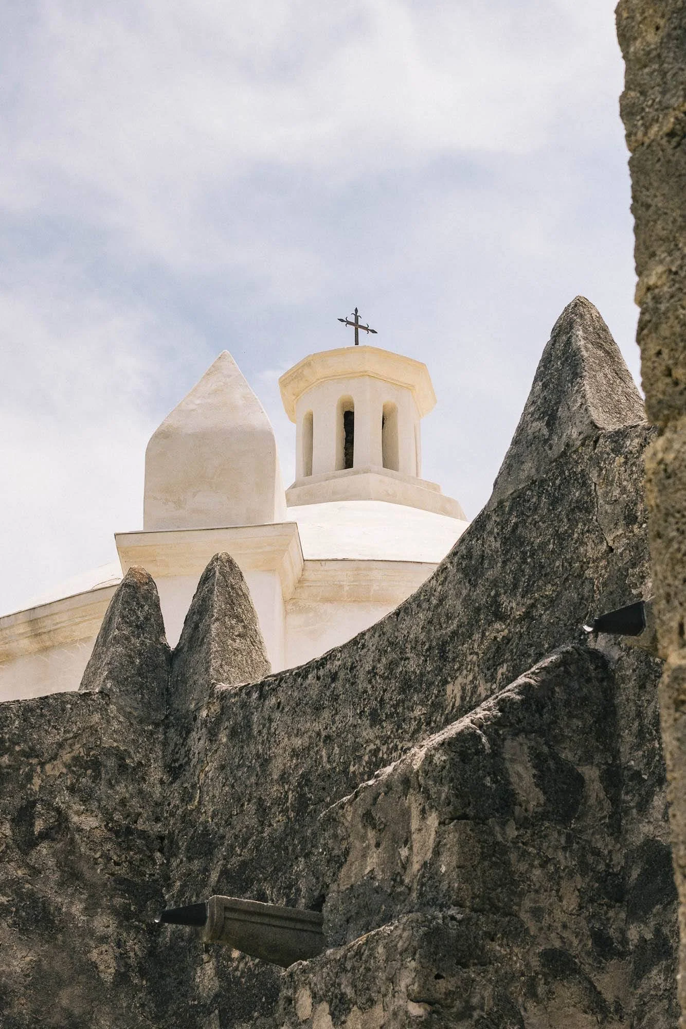 View of a white church with a cross on top, seen through dark, jagged rocks in the foreground, against a cloudy sky.