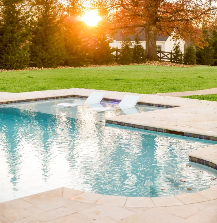 A backyard swimming pool with two white lounge chairs partially submerged in the water, surrounded by a concrete deck, with green grass, trees with autumn leaves, and a house in the background bathed in sunlight.