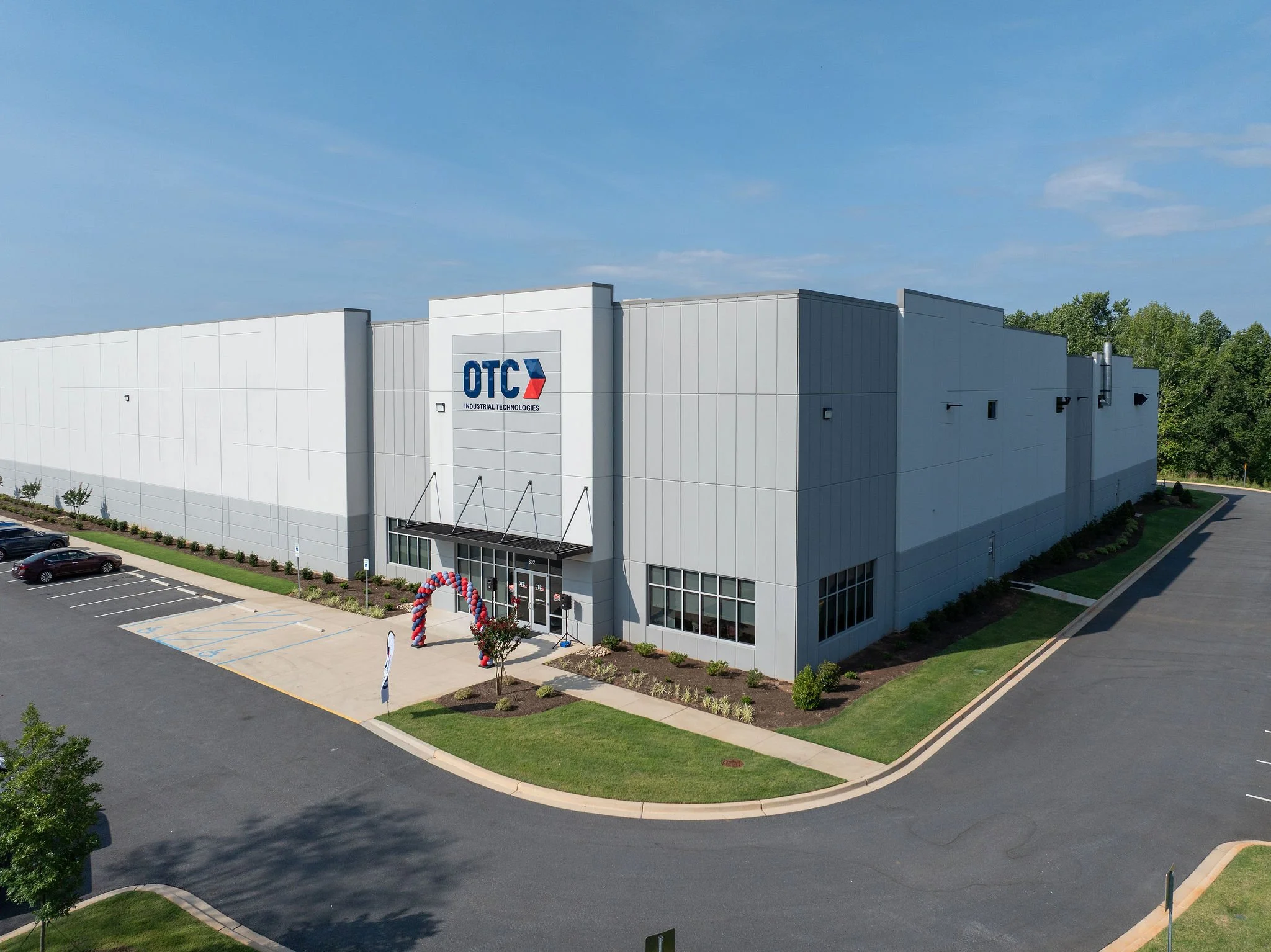 An industrial building with the logo OTIC (Industrial Technologies) on the front, surrounded by parking lot with several cars, landscaped greenery, and a blue sky overhead.