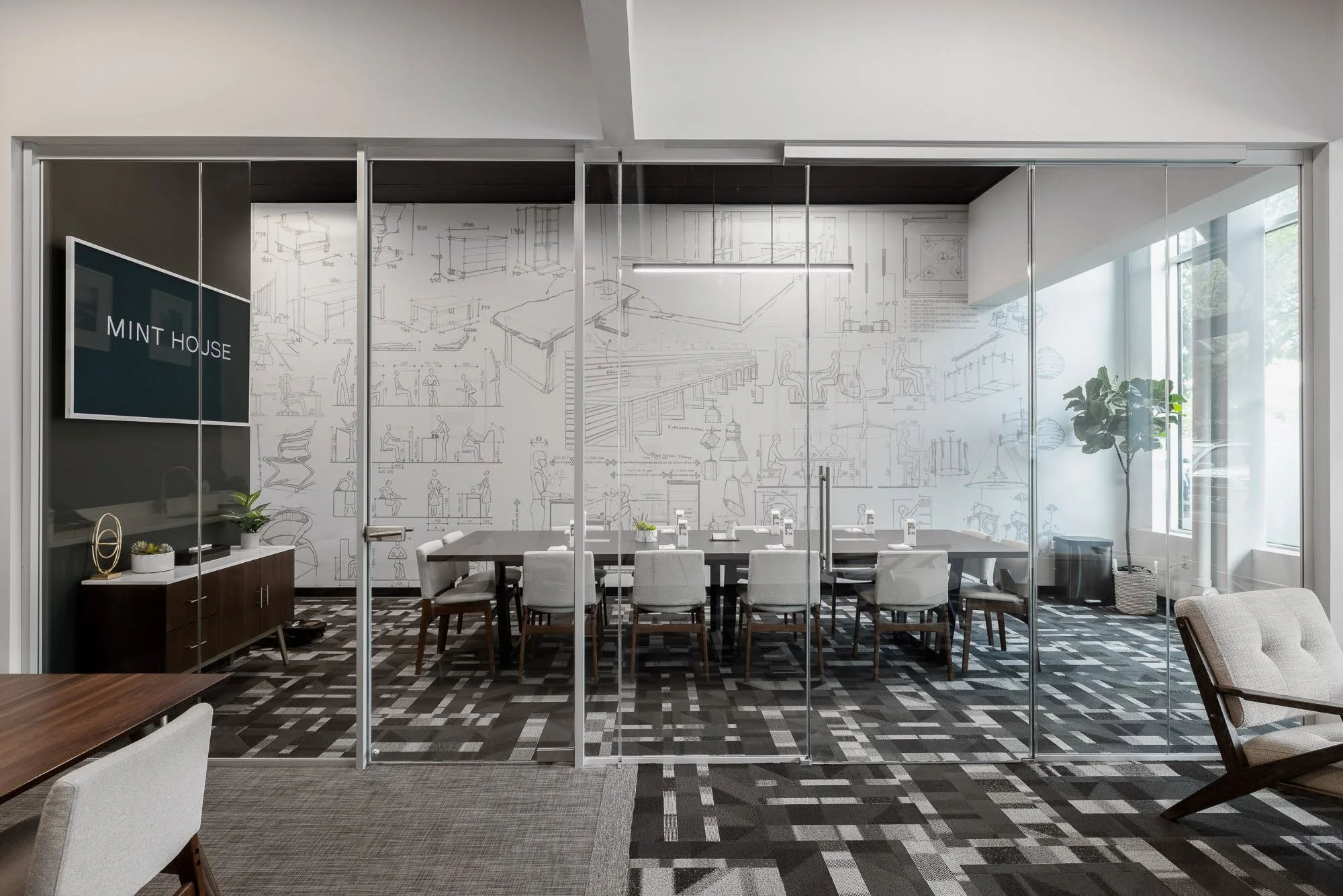 A modern conference room with a long wooden table surrounded by gray upholstered chairs. The back wall features large sketches of furniture and interior design ideas, and a black cabinet with plants and decorative items is visible on the left. The ro