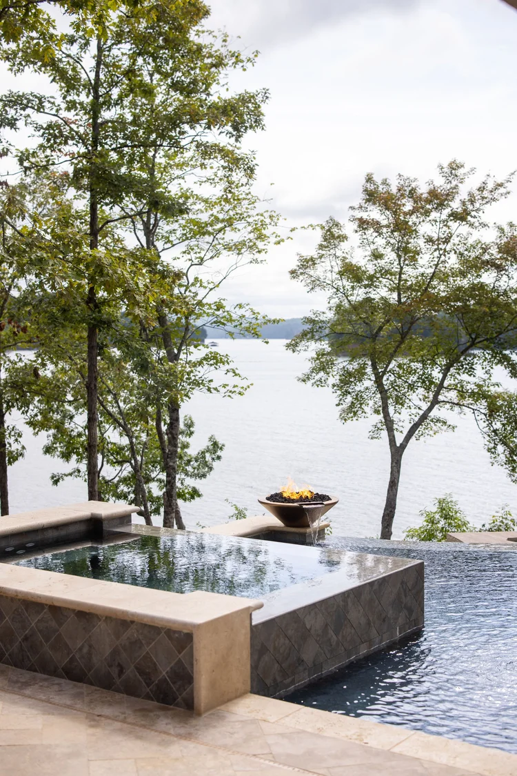 An outdoor infinity pool overlooking a lake with trees in the background, a fire feature with flames, and a cloudy sky.