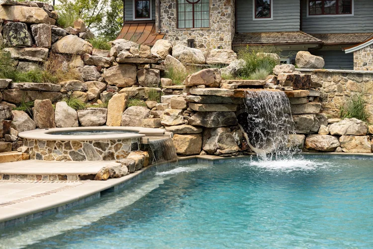 A backyard swimming pool with a waterfall feature cascading over rocks, surrounded by stone and wood structures.