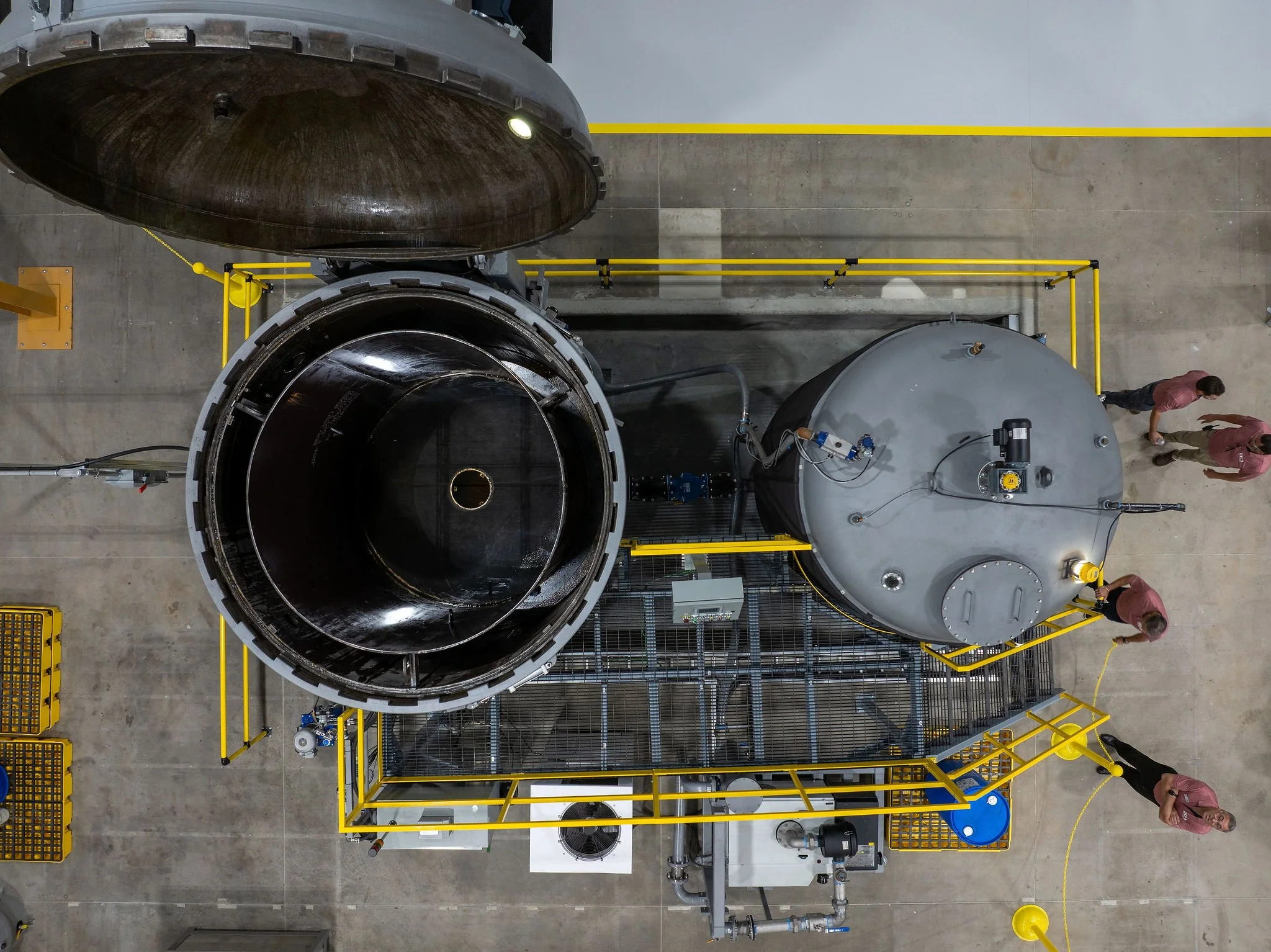 Top-down view of a large industrial machine with metallic components, pipes, and yellow safety railings, with two workers in red shirts and beige pants near the machine inside a warehouse.