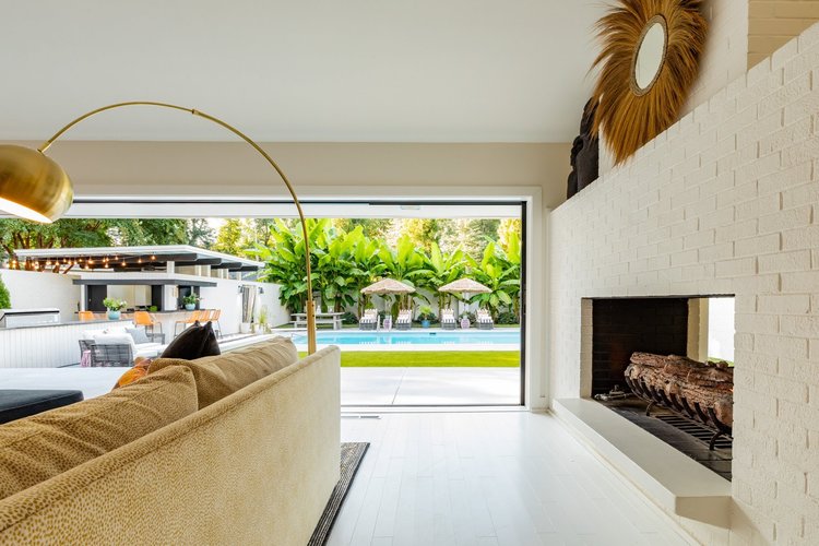 Living room with a large beige sofa, a white brick fireplace, and a sliding glass door opening to a backyard with a pool, green plants, and outdoor seating.