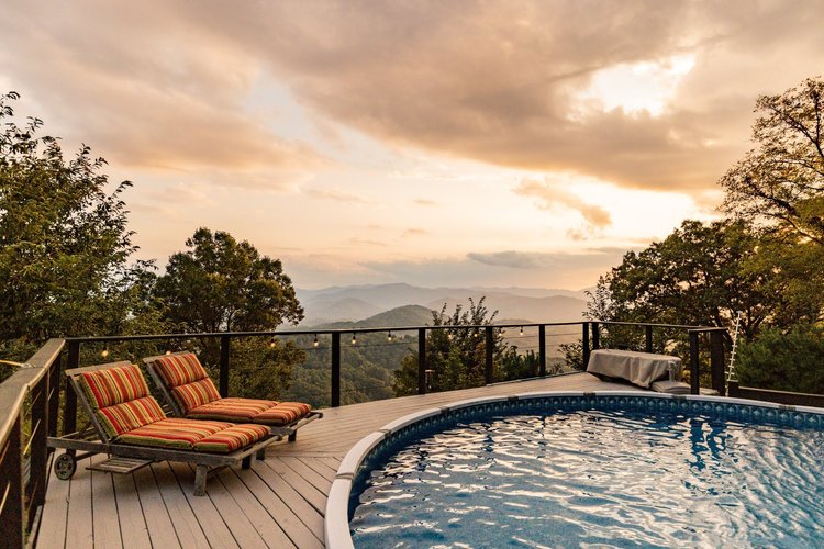An outdoor deck with a swimming pool, two lounge chairs with striped cushions, surrounded by trees and overlooking a mountain landscape at sunset.