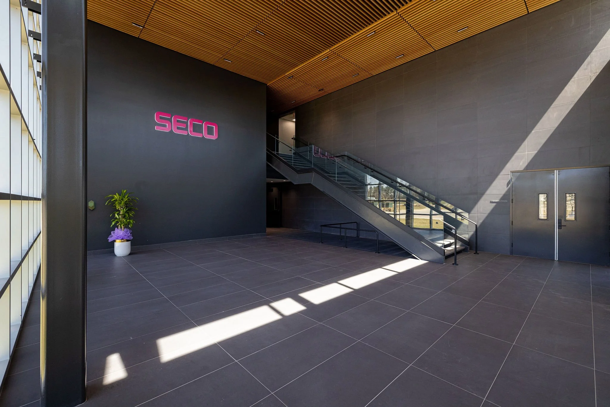 Modern building interior with dark gray walls, black tiled floors, a staircase with glass railing, a potted plant with purple ribbon, and pink 'SECO' signage.