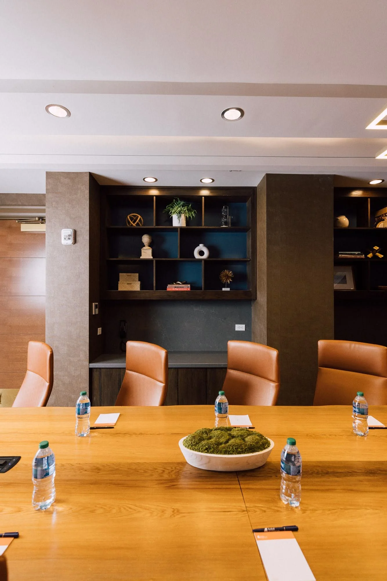 Conference room with a long wooden table, six water bottles, notepads, pens, and a bowl of moss. In the background, there are built-in shelves with decorative objects, a potted plant, and ambient lighting.