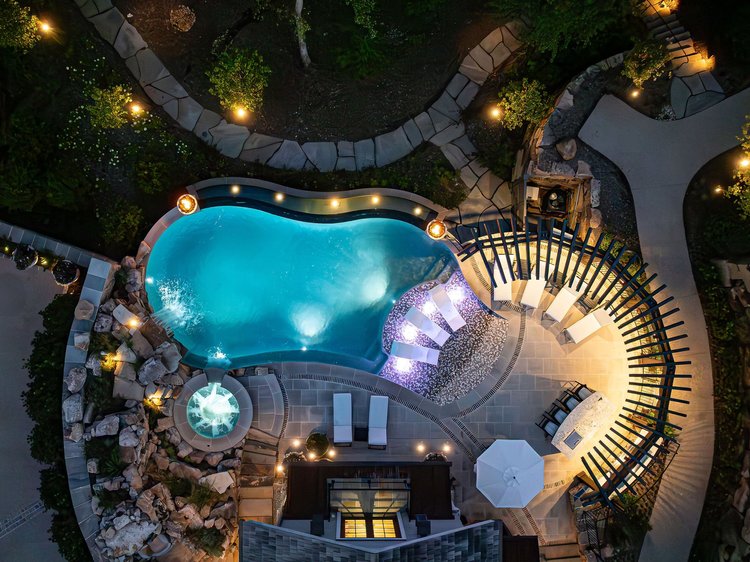 Aerial view of a backyard swimming pool area at night, featuring a kidney-shaped pool, a hot tub, outdoor seating, pathway lights, and surrounding landscaping.