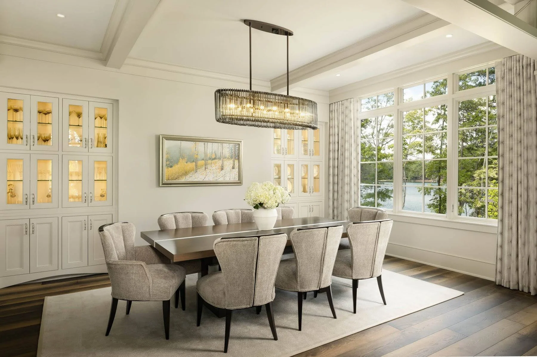Bright dining room with a wooden table, eight upholstered chairs, a white vase with flowers, large windows showing trees and a lake, built-in cabinets, and a rectangular chandelier.