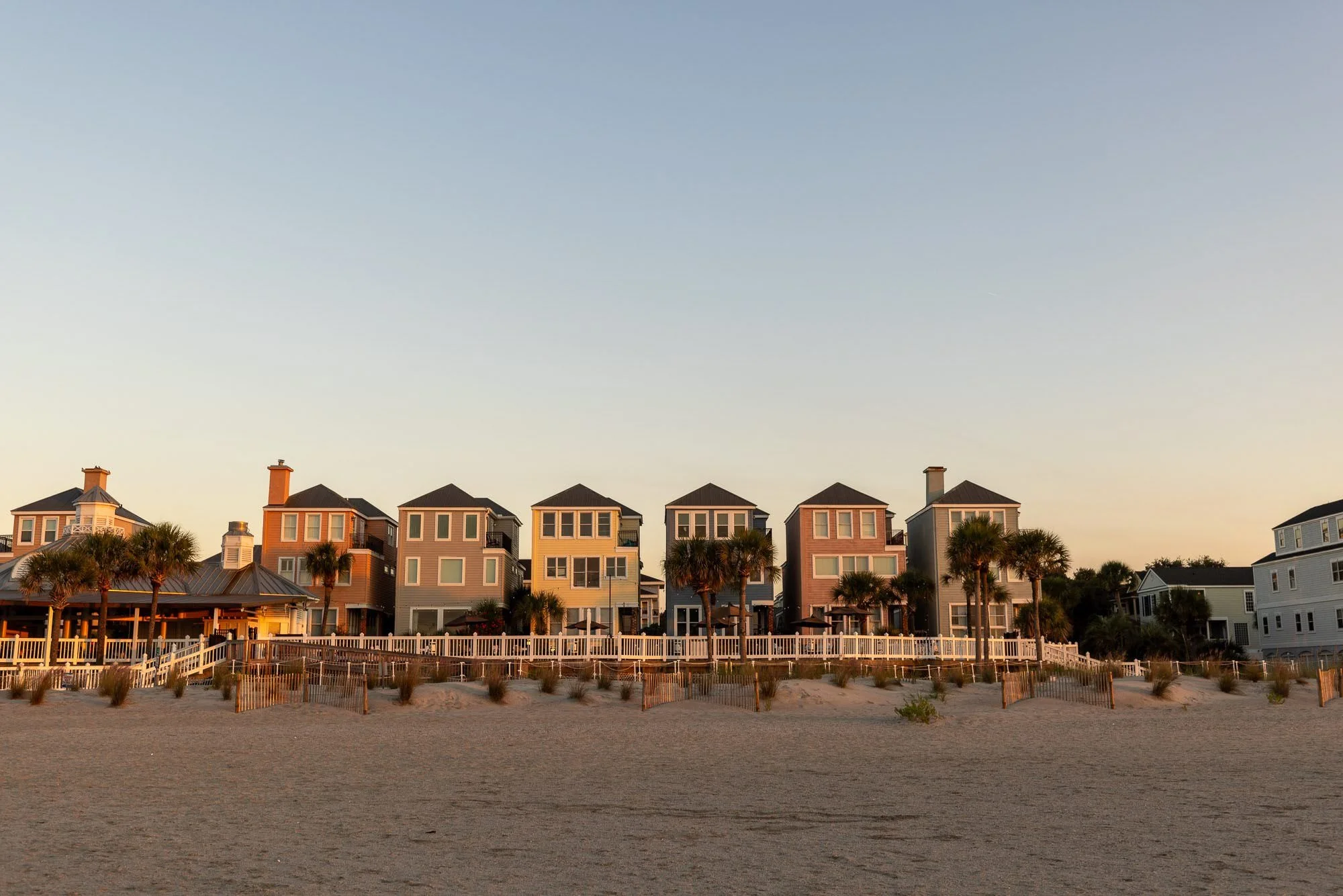 A row of colorful beach houses with palm trees in front, situated behind a sandy beach during sunset.