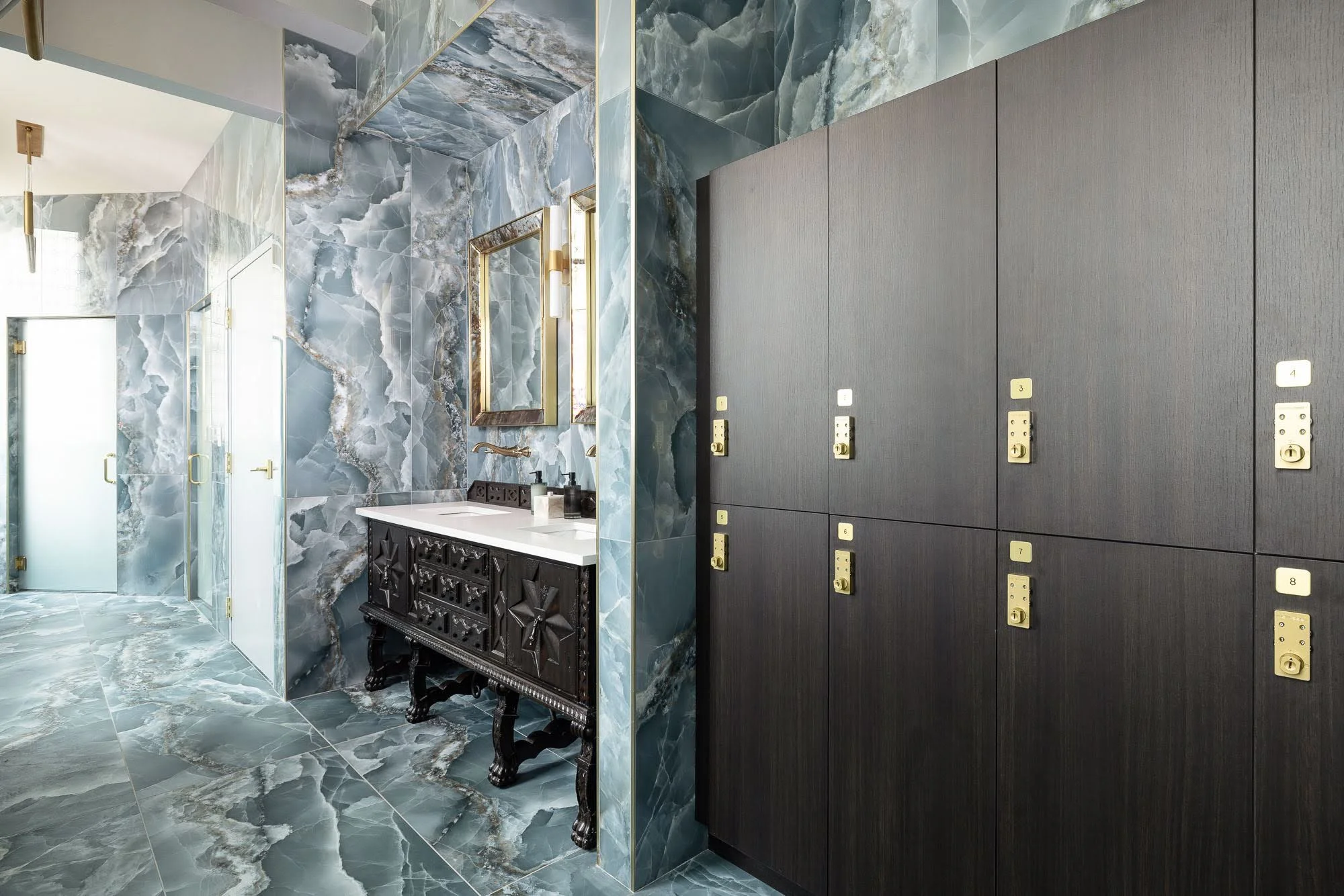 A locker area with dark gray lockers and numbered yellow labels, next to a bathroom with marble walls and floors, a black ornate vanity, a mirror, and a sink.