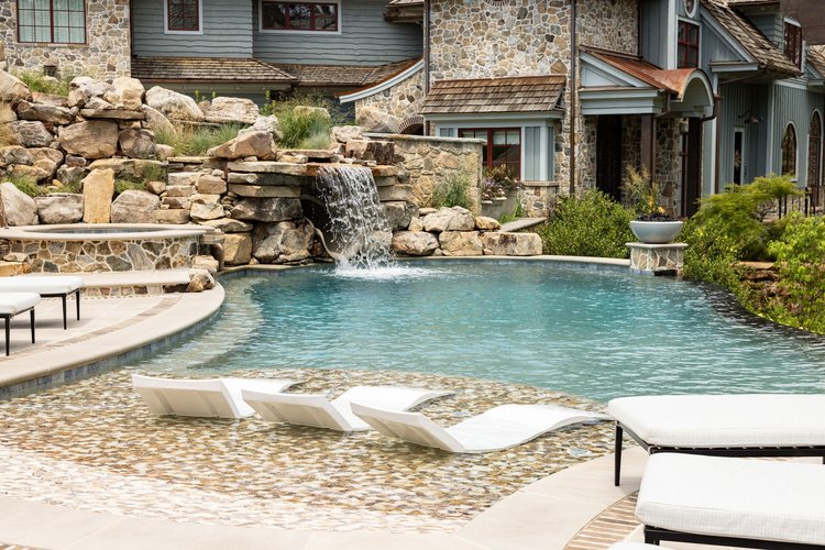 Luxury backyard with a swimming pool, waterfall feature, lounge chairs, and a stone house in the background.