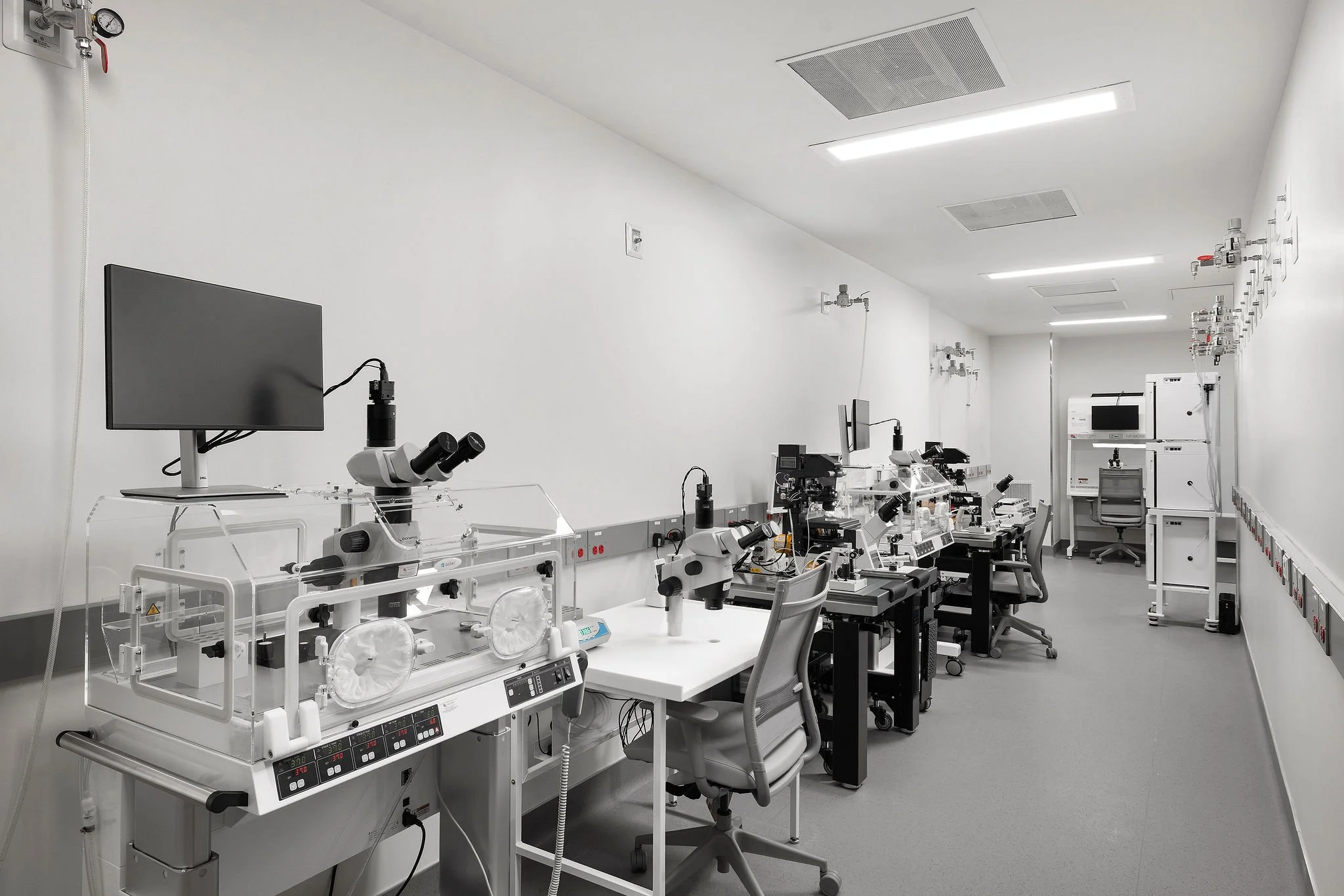 A medical laboratory with white walls and ceiling. Contains various medical equipment, microscopes, monitors, and workstations arranged neatly. Fluorescent lights and ceiling vents are visible.