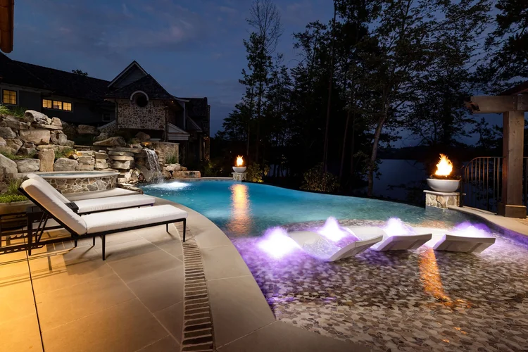 A nighttime view of an illuminated backyard swimming pool with submerged lounge chairs, fire features, a cascading waterfall, stone landscaping, and nearby trees overlooking a lake.