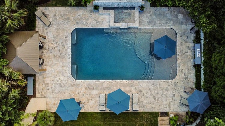 Aerial view of a backyard swimming pool with three blue umbrellas, lounge chairs, surrounding lush green trees, and stone paving.