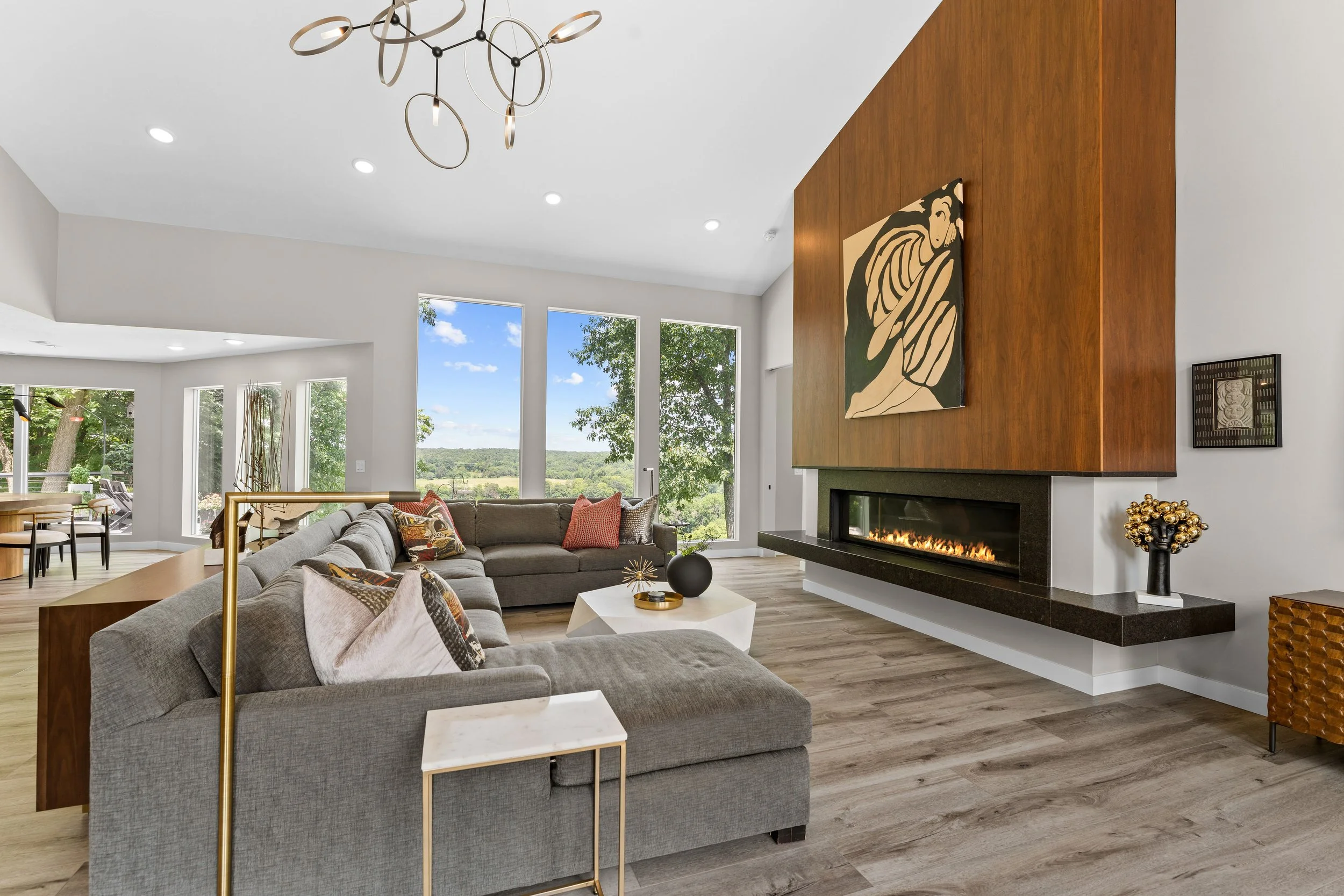 Living room with a stone fireplace, built-in wooden shelves, a ceiling fan, large windows with curtains, and furniture including armchairs, a sofa, and a coffee table.