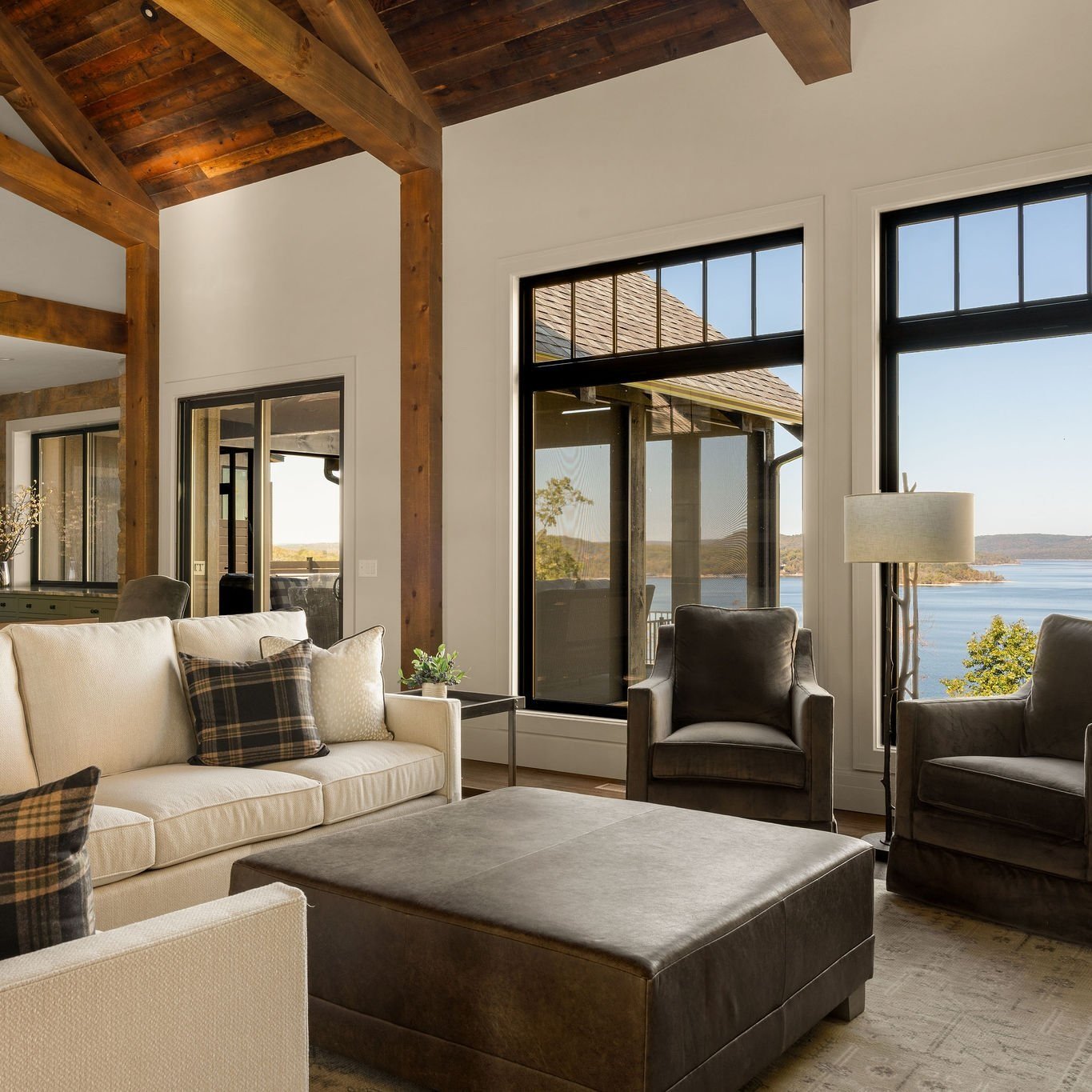 Living room with large windows showing lake view, beige and dark gray seating, wooden beams ceiling, and a view of the outdoors.