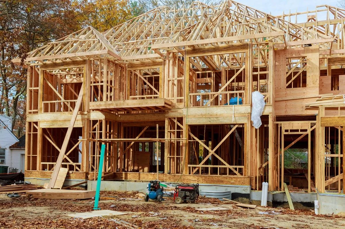 Wooden framing structure of a house under construction with scaffolding, construction tools, and building materials on ground.