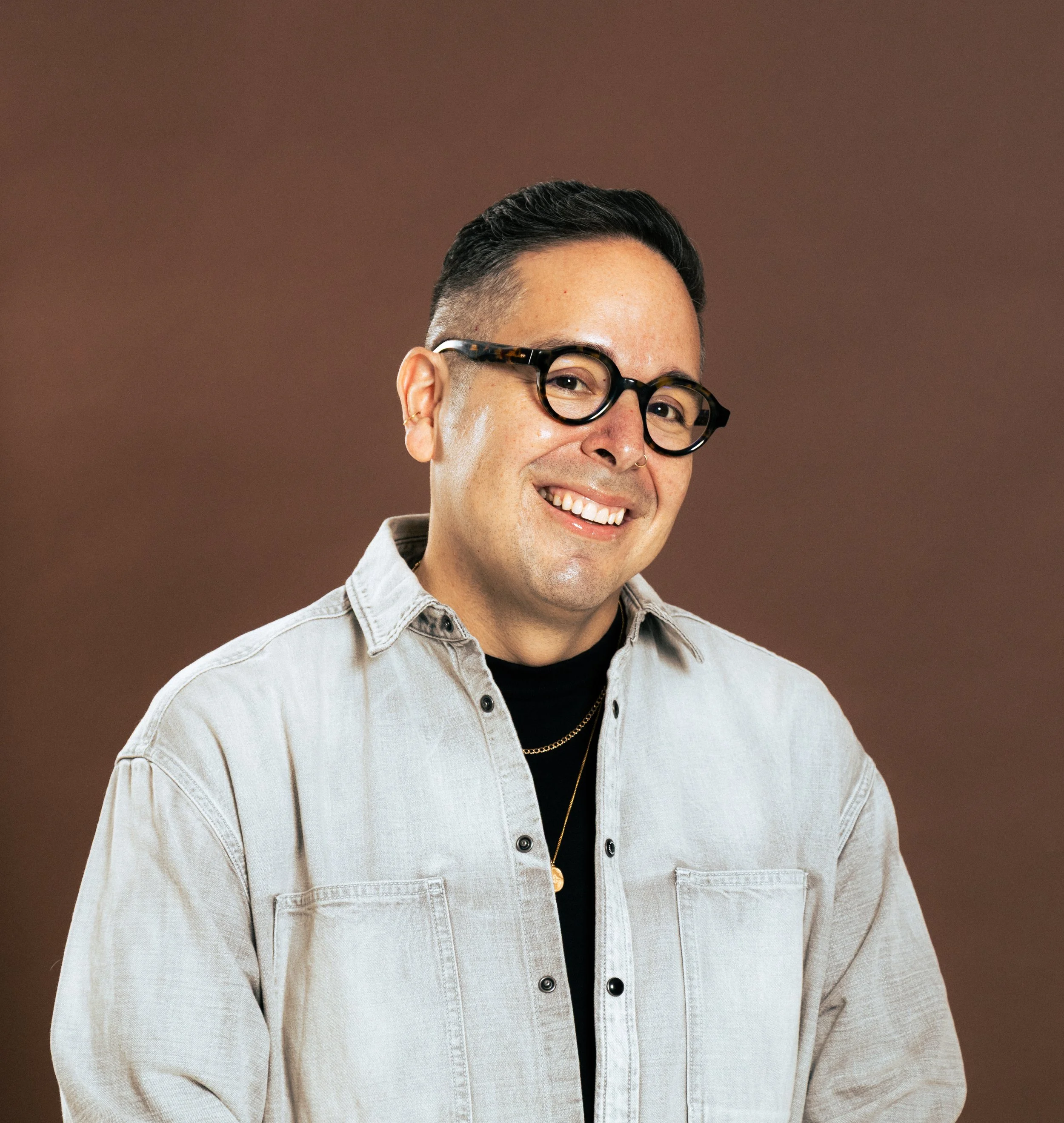 A smiling man with glasses, short dark hair, wearing a light-colored denim shirt and layered necklaces, against a plain brown background.