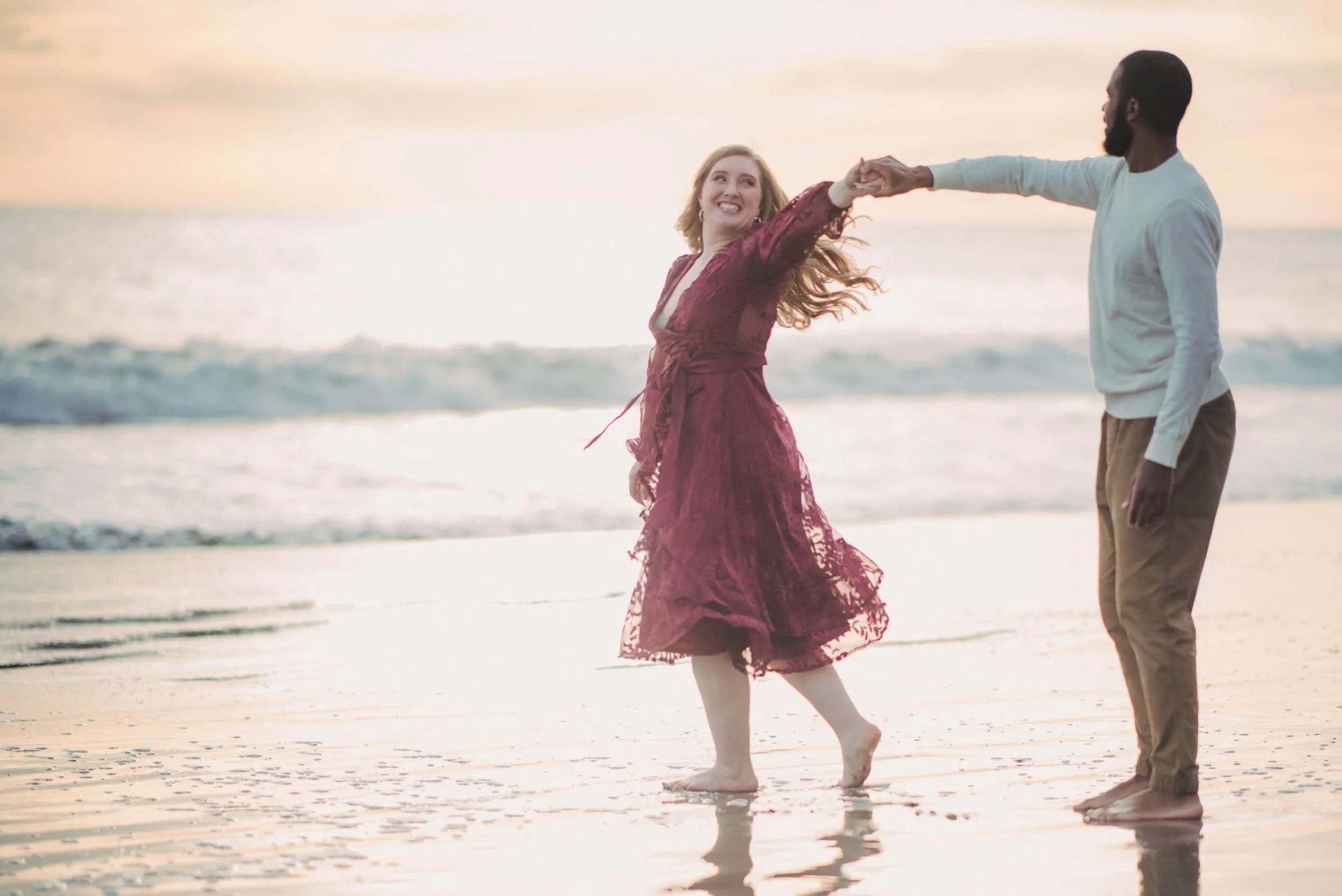 A happy couple dancing on a beach at sunset, with the woman in a burgundy lace dress and the man in a white long-sleeve shirt and khakis.