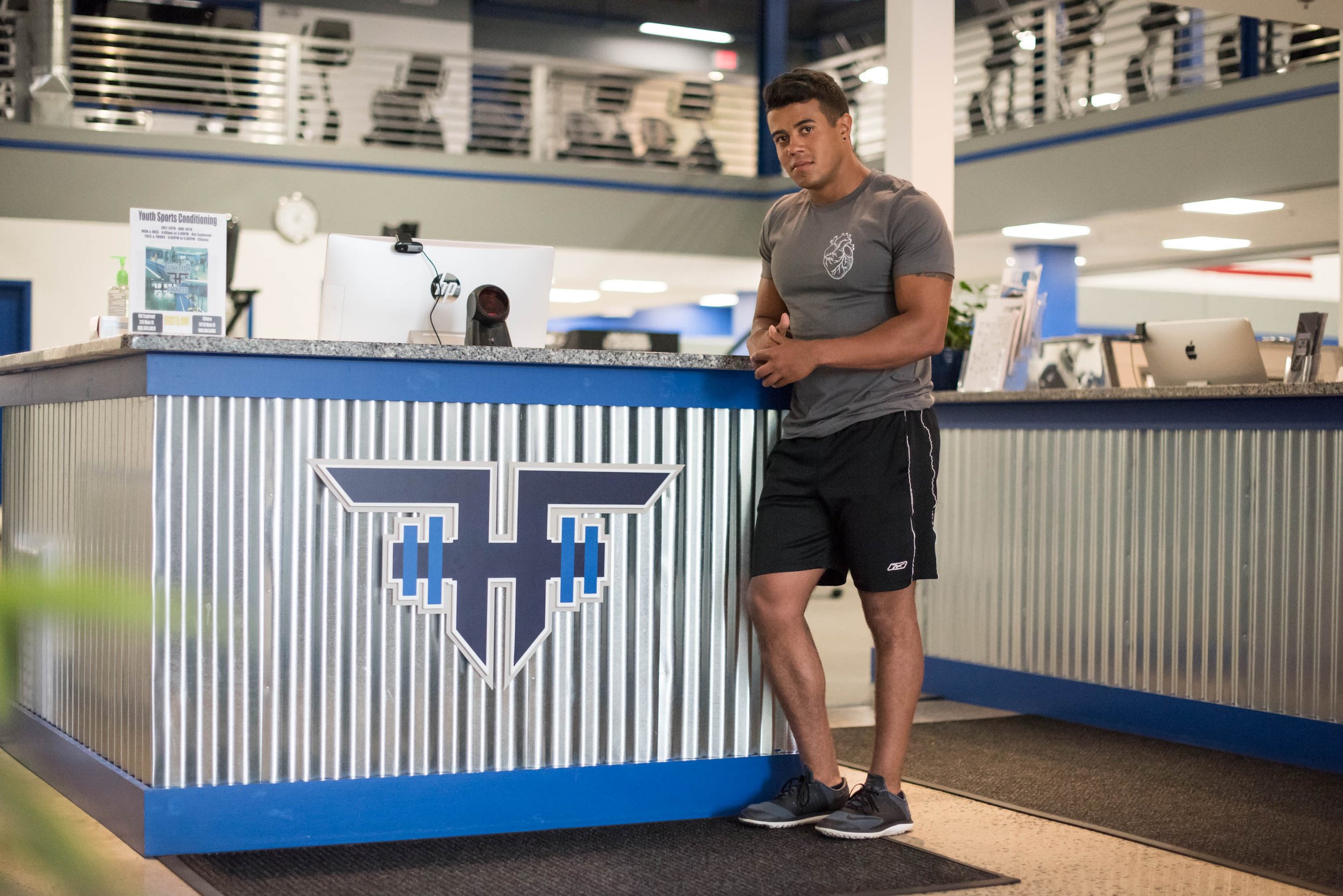 A person standing casually at a metallic and blue gym reception desk, with gym equipment visible in the background.