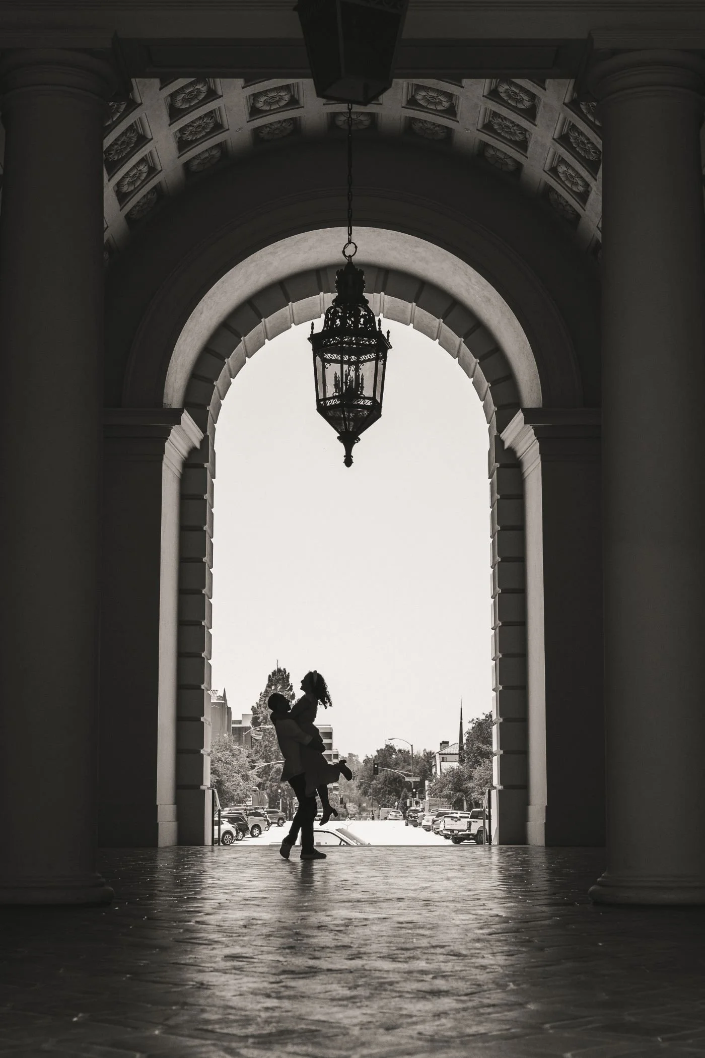 A silhouetted couple, with one person lifting the other, standing under a large archway with columns, a hanging lantern, and a street scene in the background.