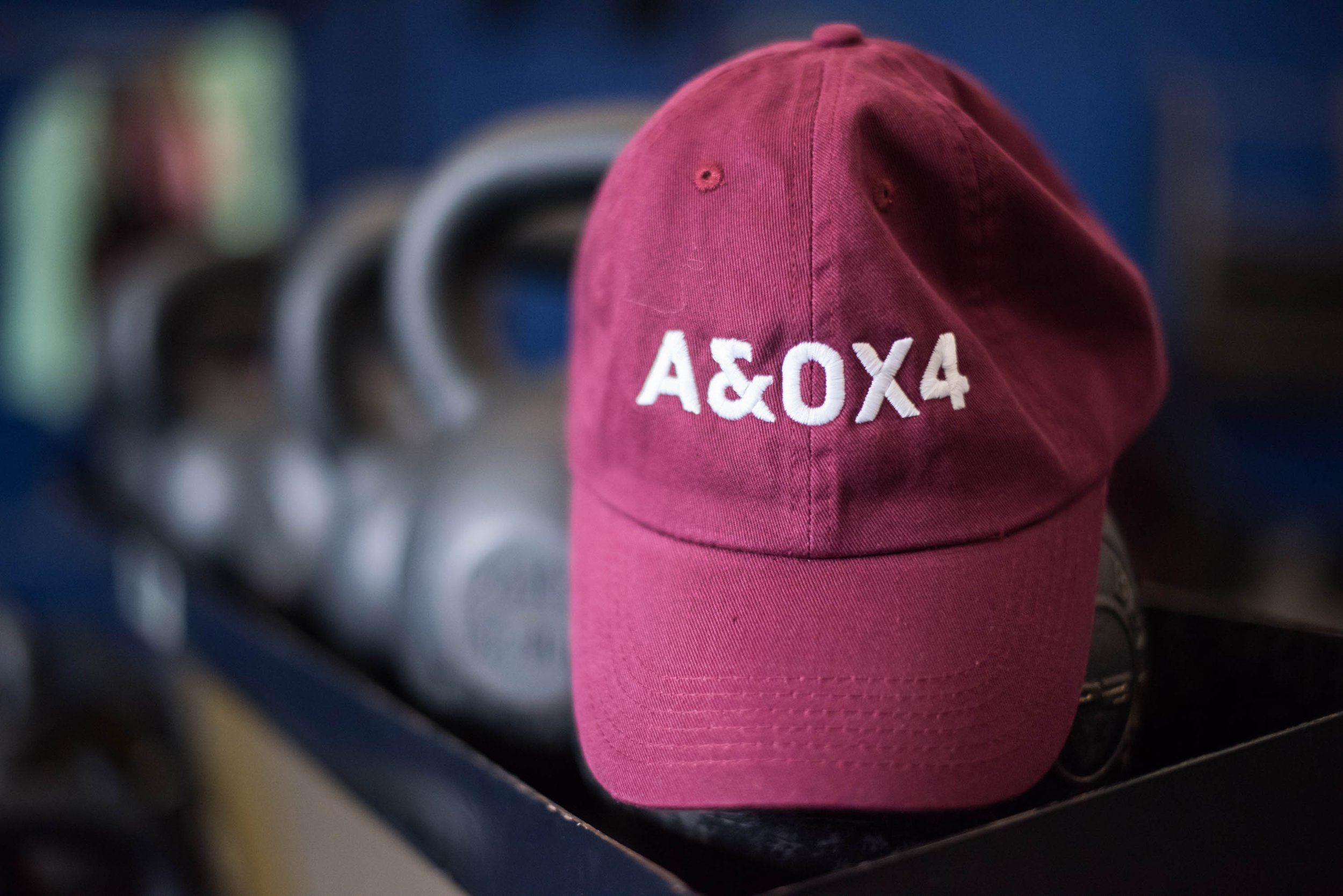 Maroon baseball cap with "A&OX4" text in white, placed on a gym rack with kettlebells in the background.