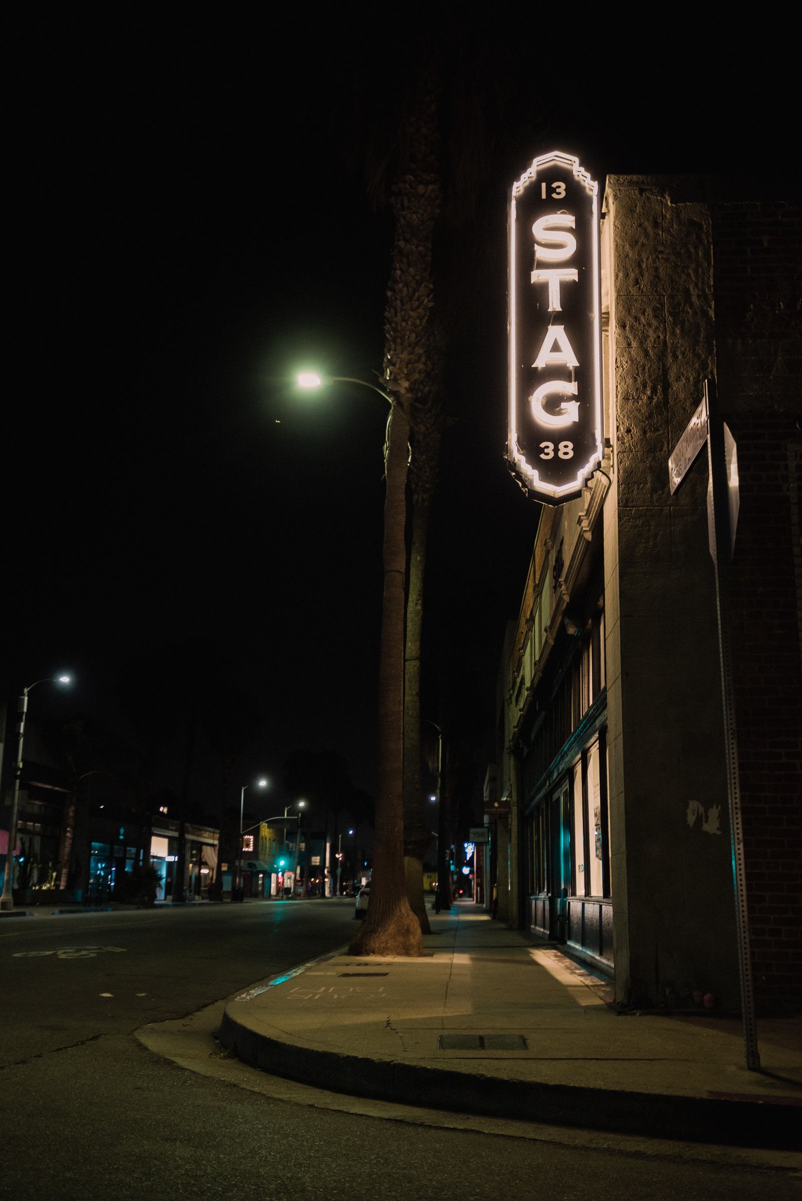 Night street scene with an illuminated "STAG" sign on a building, featuring a dimly lit sidewalk, palm tree, and street lamps.