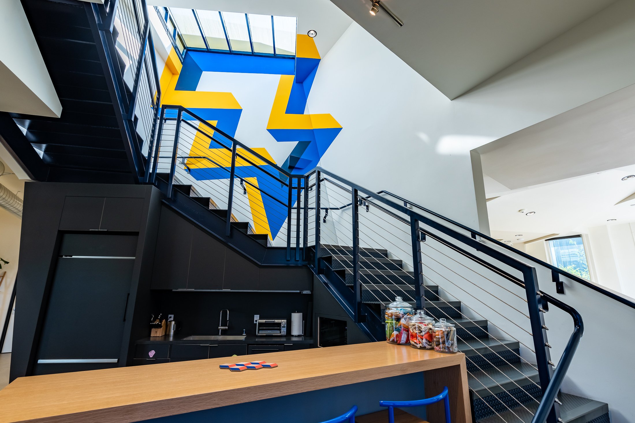 Modern interior with a black metal staircase, colorful geometric wall art in blue and yellow, and a small kitchen area. A wooden counter with jars of snacks is visible in the foreground.Media.Monks los Angeles - commercial interior photography