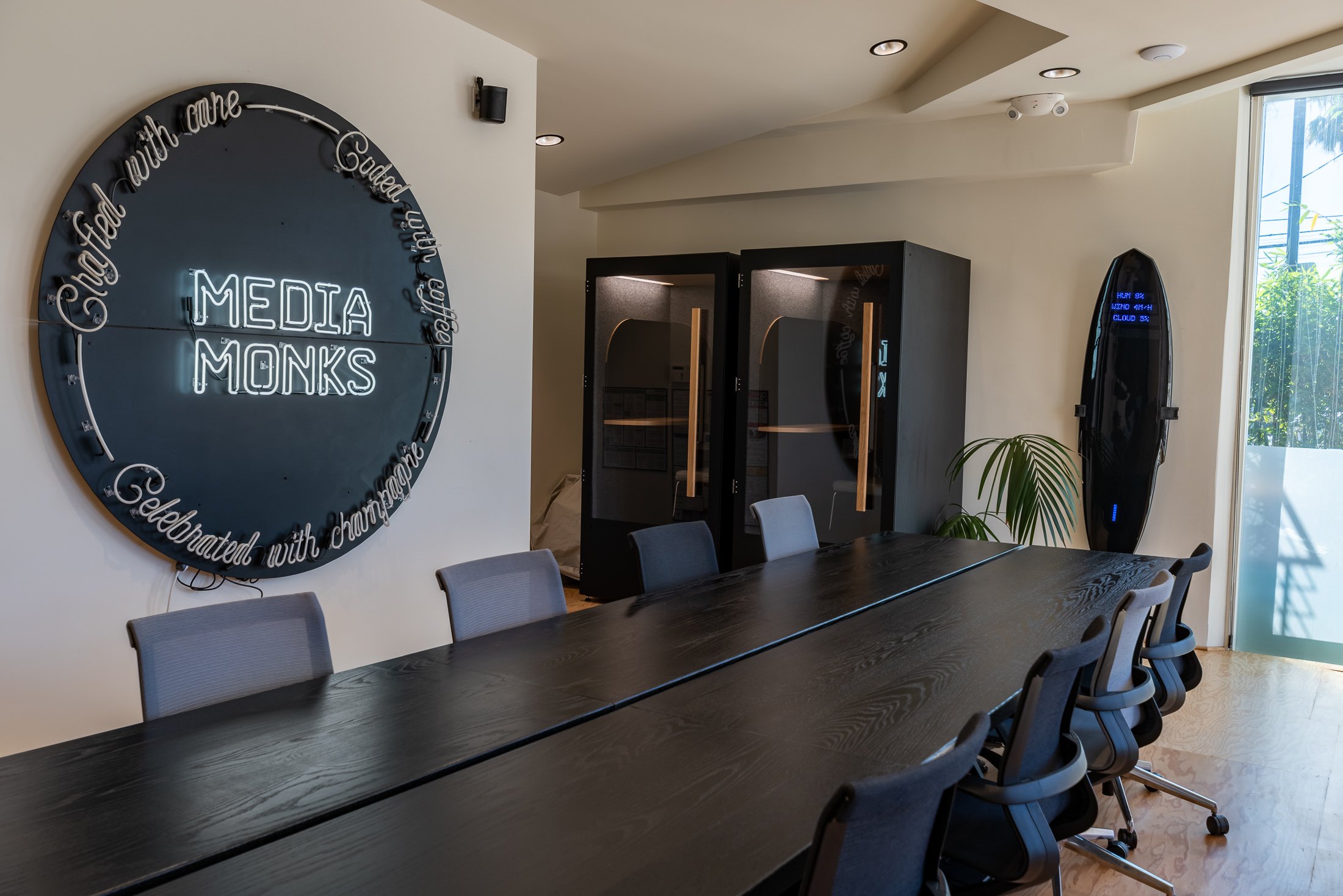 Modern office meeting room with a long black table, gray chairs, a neon sign on the wall displaying 'Media Monks,' phone booths, a plant, and a surfboard-shaped digital display. Media.Monks los Angeles - commercial interior photography
