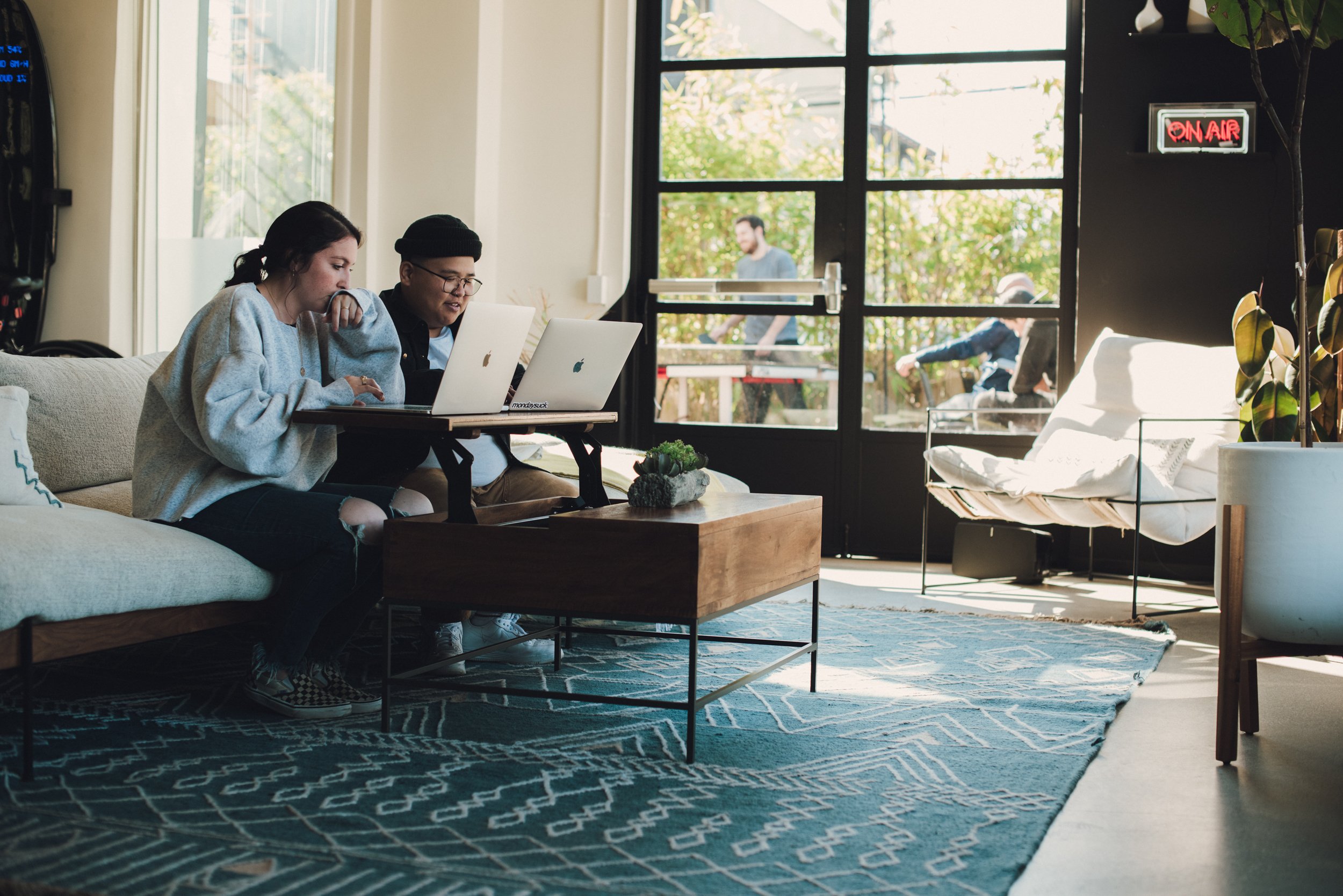 Two people sitting on a couch in a well-lit modern living room or office space working on laptops, with a large window in the background and a decorative rug on the floor. Media.Monks los Angeles - commercial interior photography