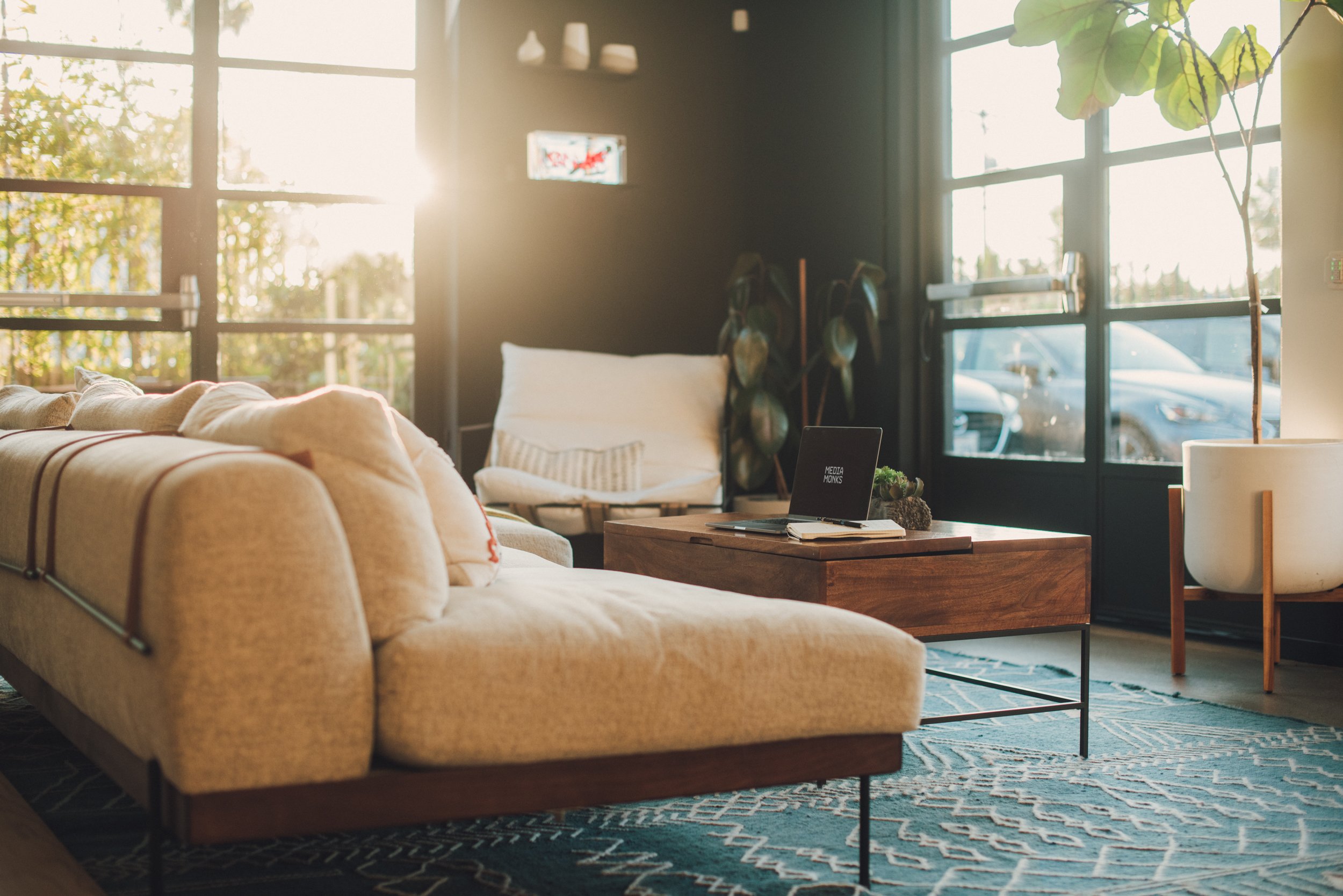 Modern living room with a beige sofa, wooden coffee table, and large windows letting in sunlight. A laptop sits on the table with a potted plant. A patterned rug covers the floor, with a white armchair and plant visible in the background.