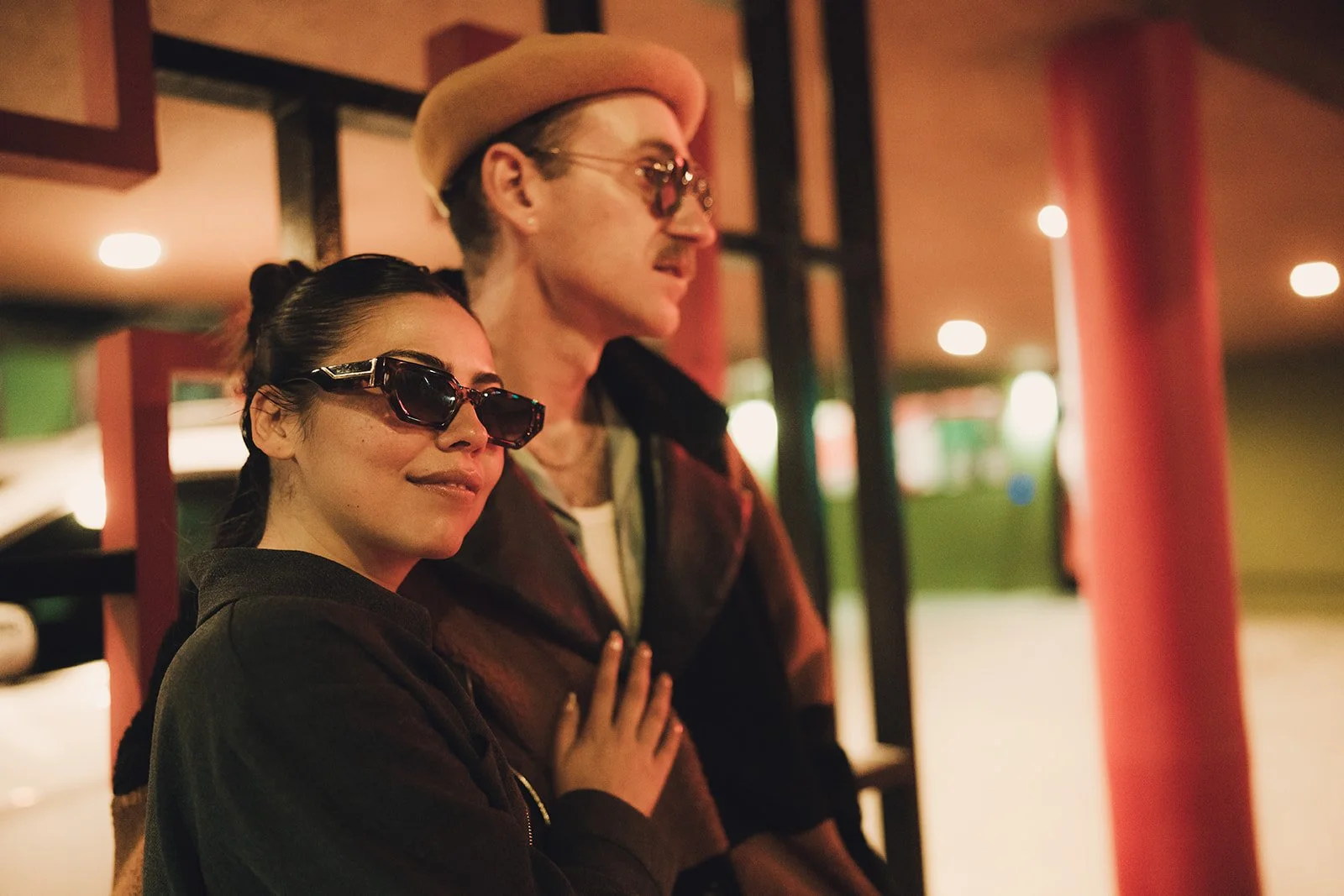 A stylish woman and man wearing sunglasses, standing together indoors, with soft lighting and blurred background ambiance.
