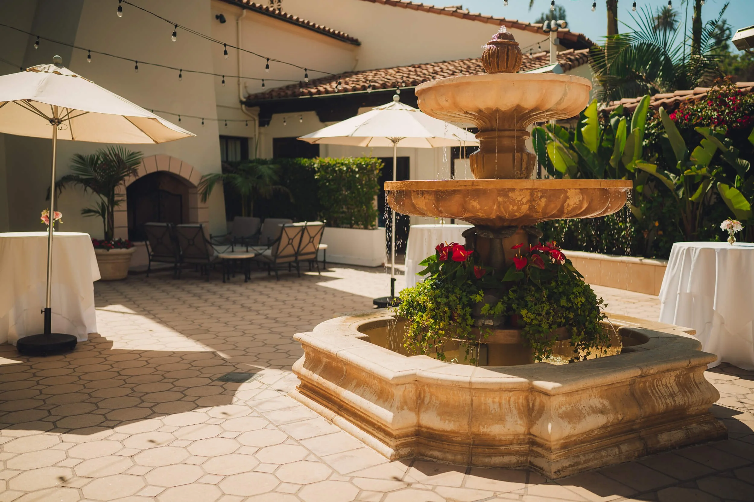 A courtyard featuring a large ornate stone fountain with water cascading from its tiers, decorated with flowers and surrounded by outdoor tables, umbrellas, and lush greenery.