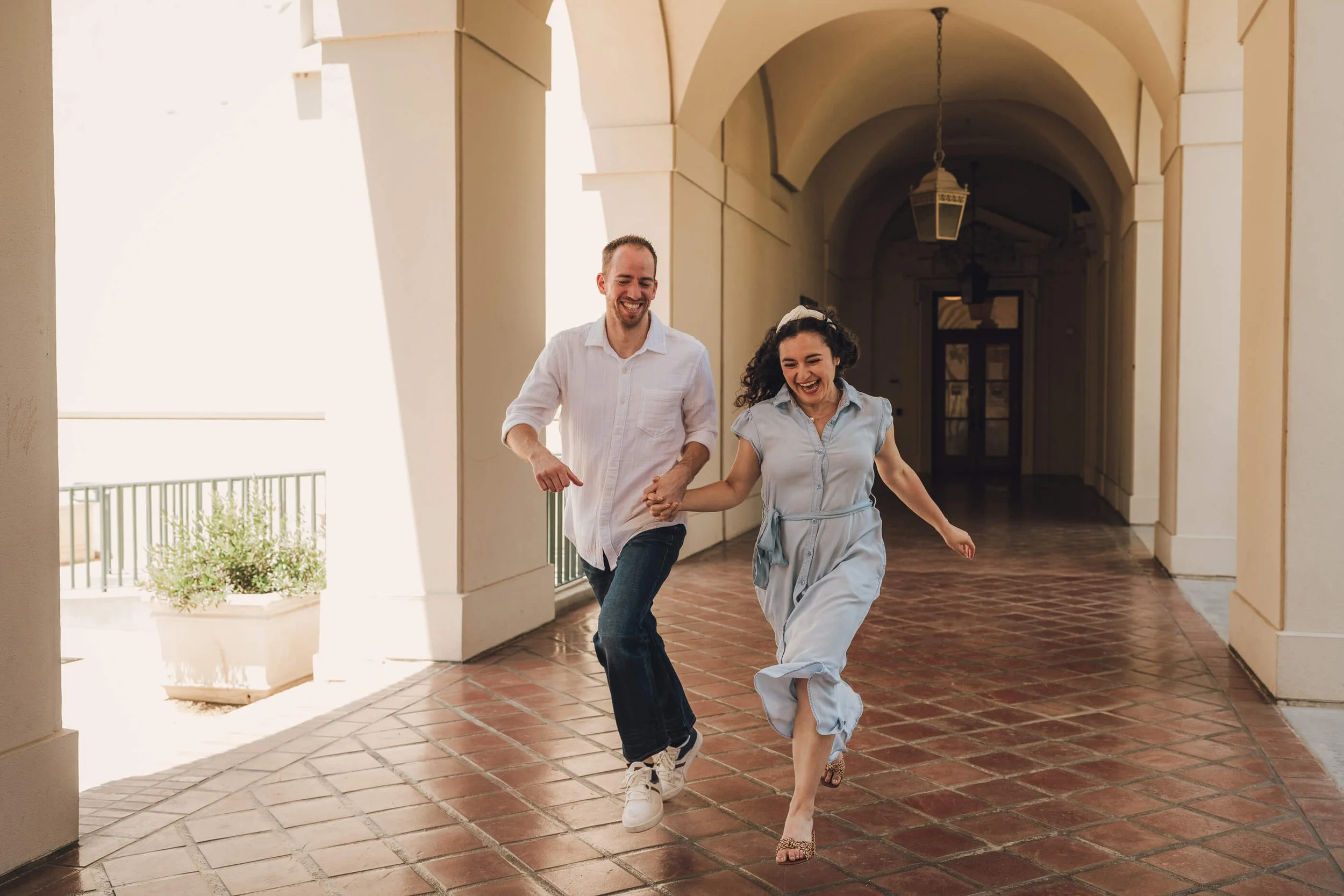 A happy man and woman running hand in hand through a covered outdoor corridor with arches and tiled floor.