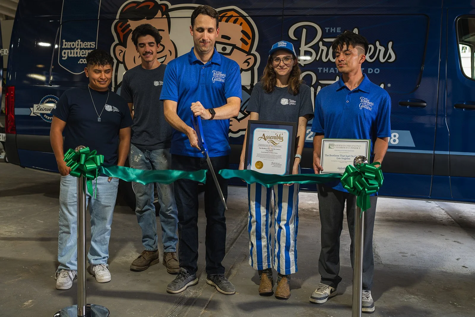 Five people standing behind a green ribbon with two holding framed certificates, at a ribbon-cutting event for Brothers Gutter company, in front of a branded van.