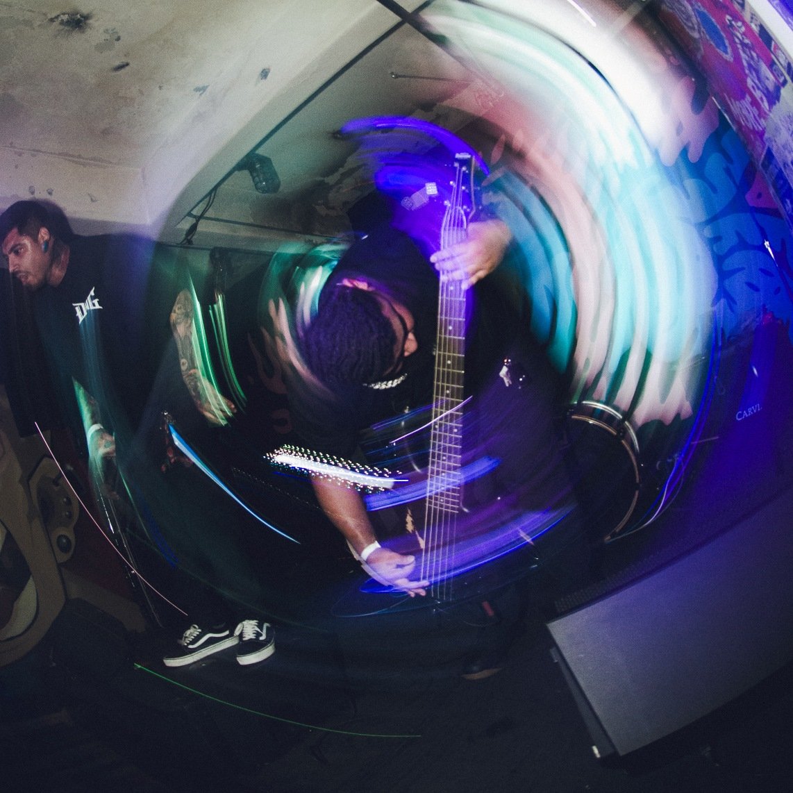 Musicians performing in a dimly lit setting with a swirling motion blur effect, emphasizing the movement and energy. One musician plays a guitar or bass, while another stands nearby. The background features vibrant graffiti art.