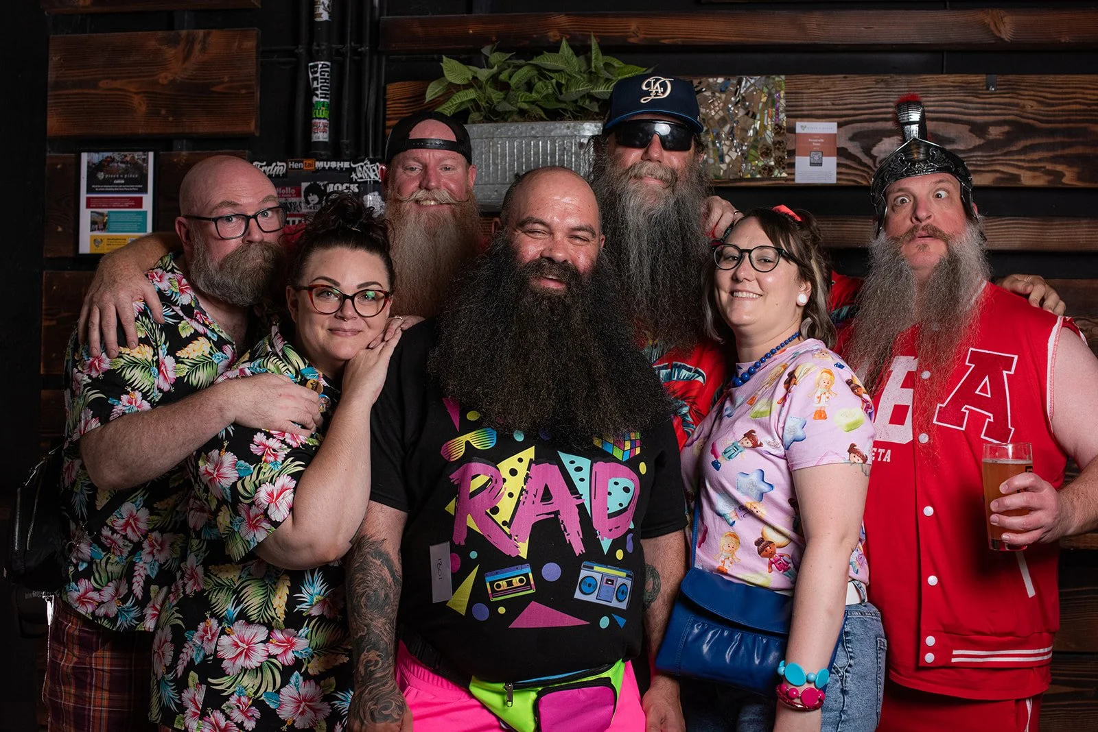 Group of seven people with beards, glasses, and colorful clothing indoors, smiling and posing for a photo.