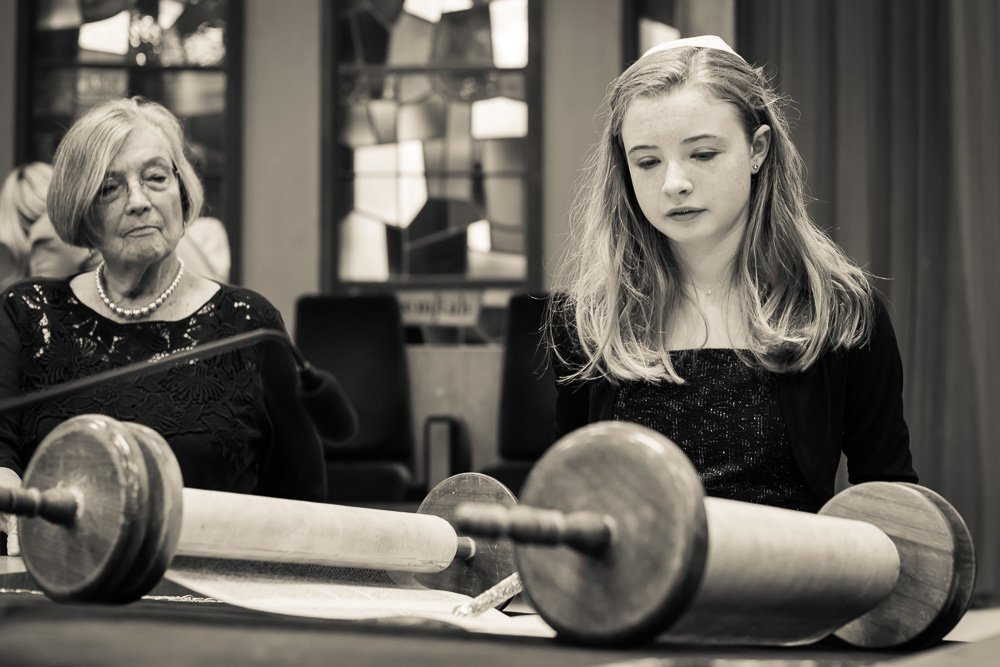 A young girl and an older woman sitting at a table with a Torah scroll in front of them in a synagogue or religious setting.