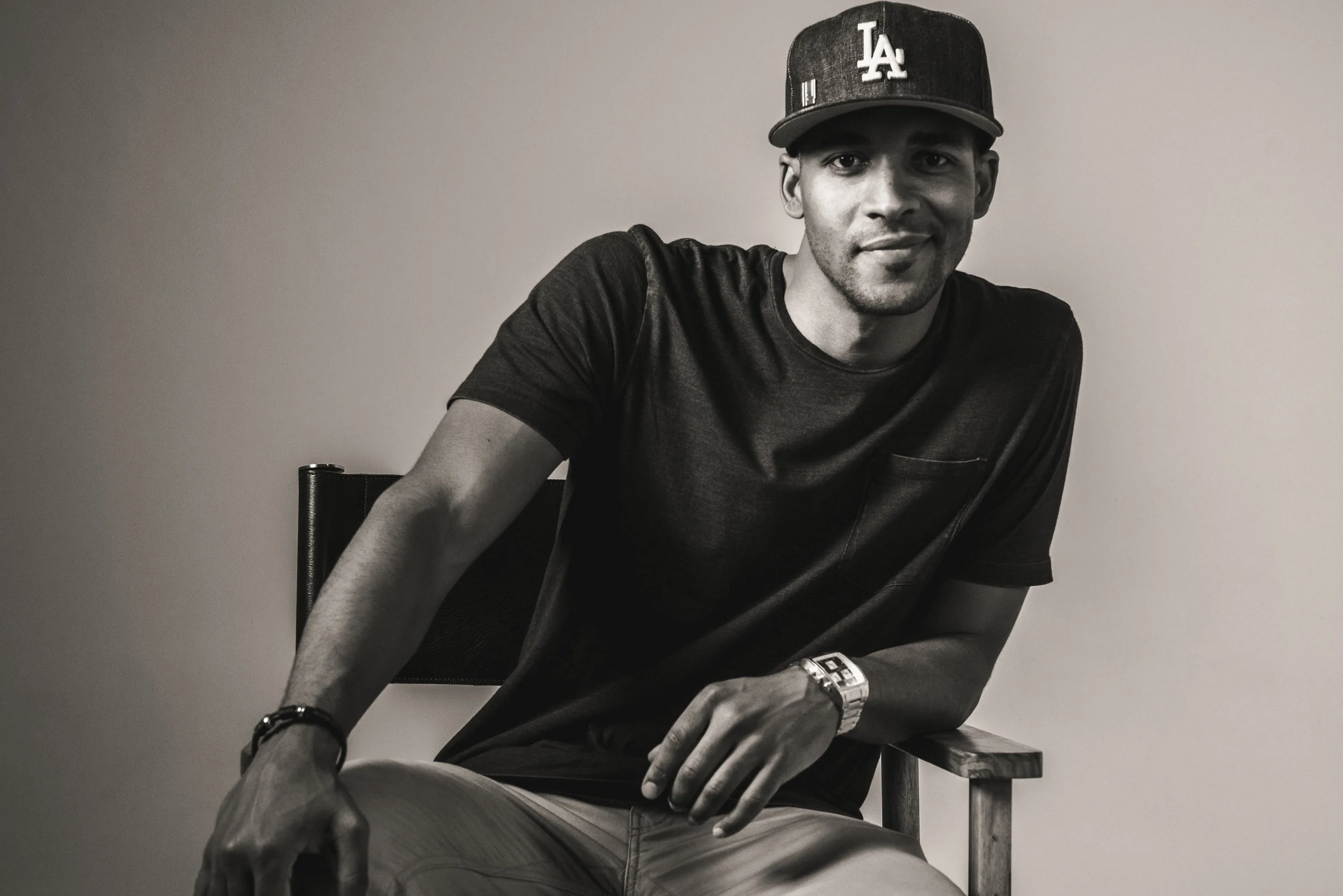 A man sitting on a chair, wearing a baseball cap with 'LA' logo, a black t-shirt, a wristwatch, and bracelets on his left wrist, looking at the camera with a slight smile.