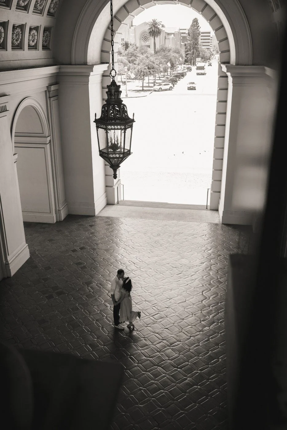 A black-and-white photo of a couple dancing in a grand archway with a chandelier hanging above them, overlooking a sunny street with parked cars and trees.