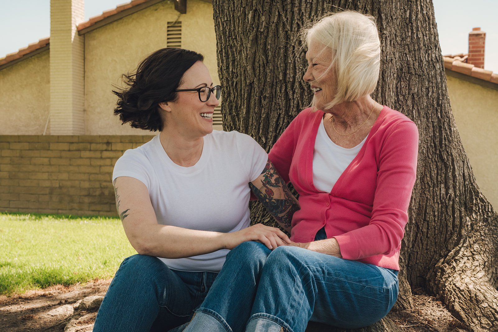 Two women sitting and laughing near a tree, one with short hair and glasses, the other older with white hair wearing a pink cardigan.
