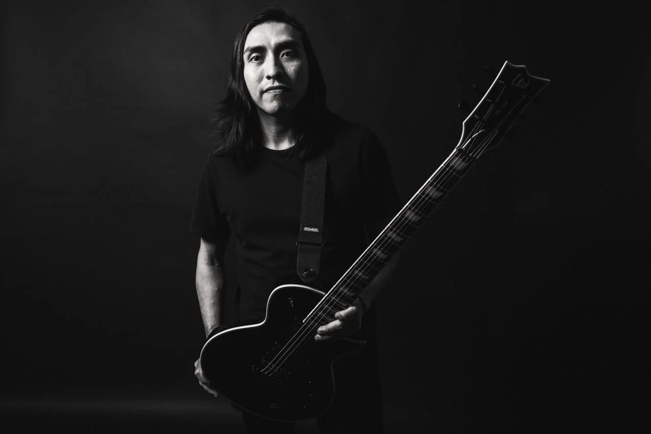 A man with long dark hair and a serious expression holding an electric guitar in a dark room, black and white photography.