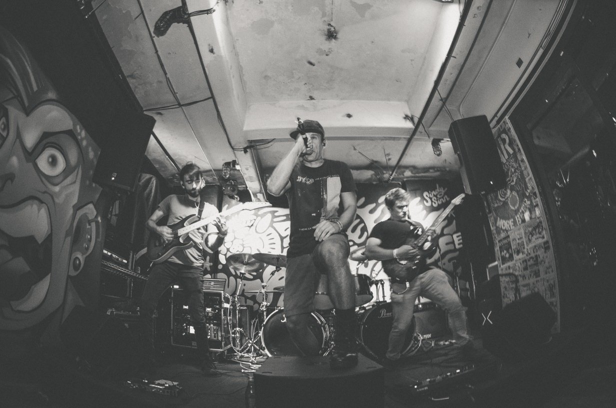 Black and white photo of a rock band performing on stage with a microphone, guitars, and drums. The stage has graffiti art in the background and overhead lighting.