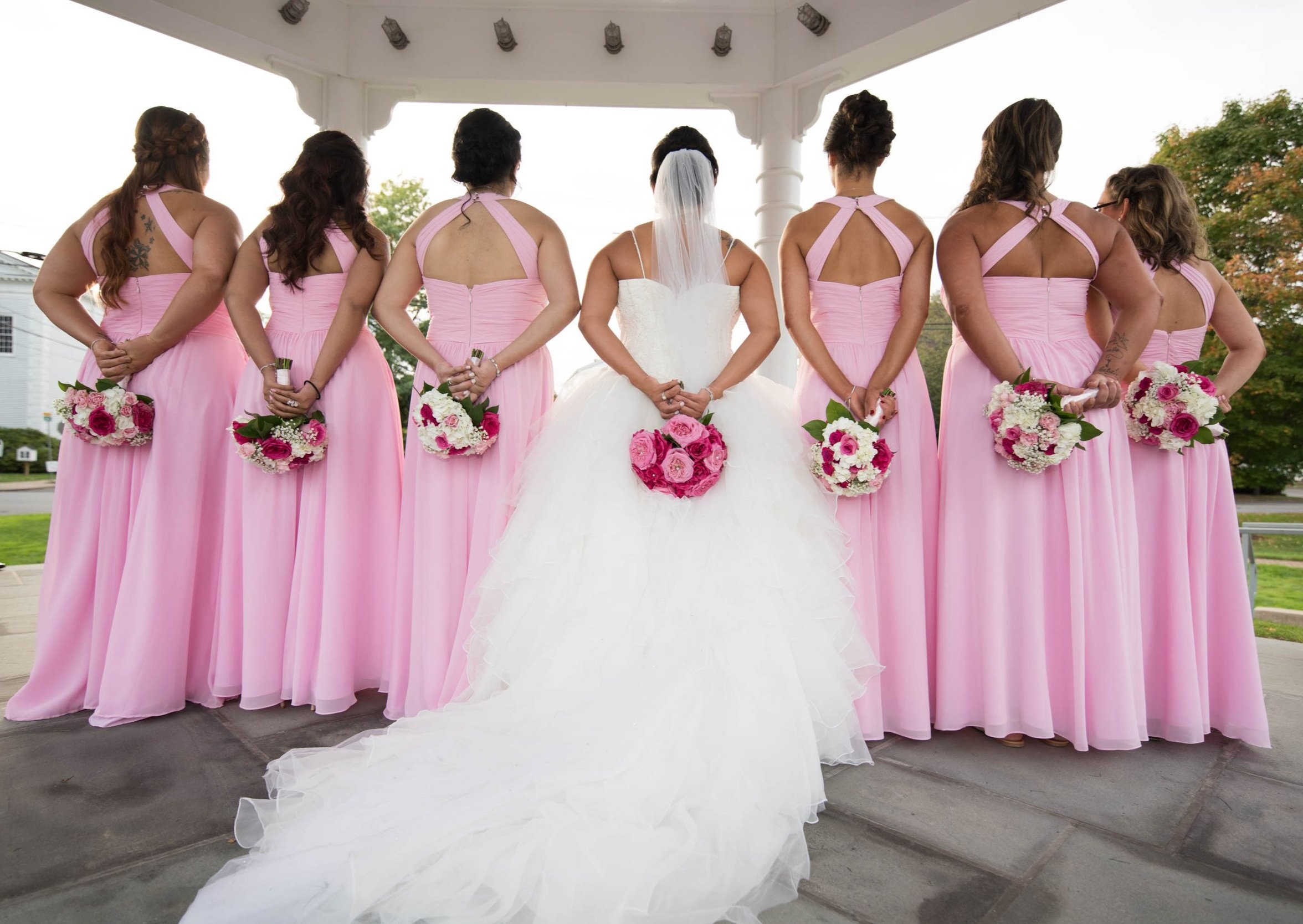 Bride and bridesmaids in pink dresses holding bouquets, standing in a gazebo.
