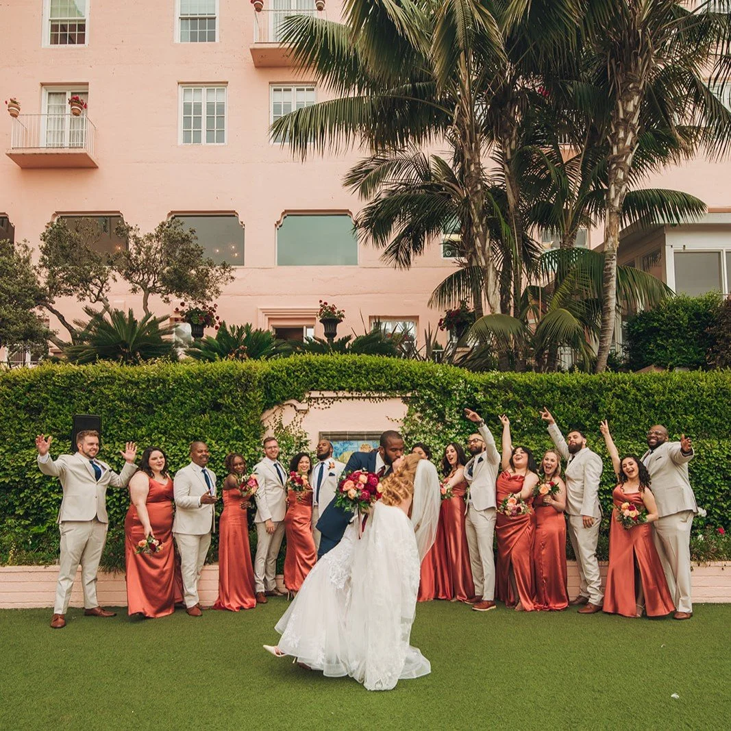 A wedding celebration with a bride and groom kissing in front of a group of wedding party members, who are dressed in peach and beige attire, outdoors with lush greenery, palm trees, and a pink building in the background.