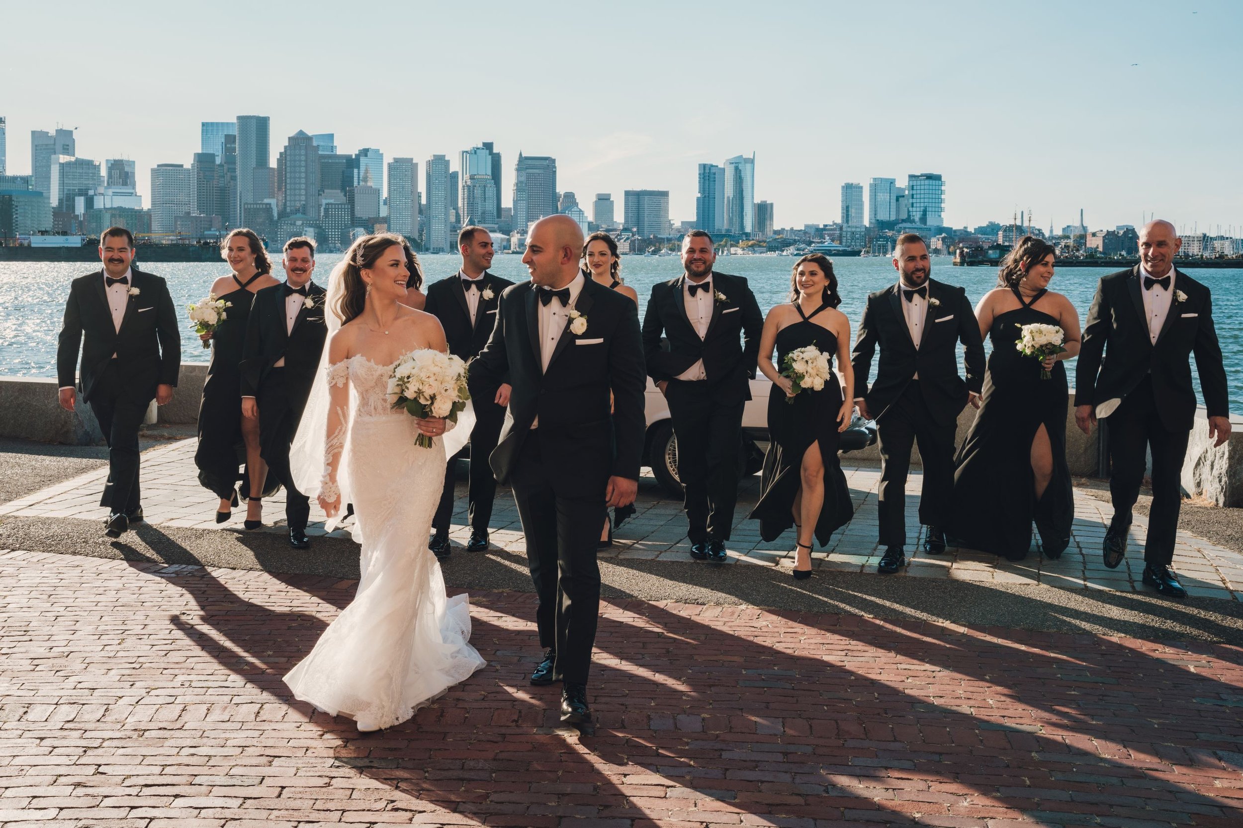 A wedding party poses outdoors by a waterfront with a city skyline in the background. The group includes a bride in a white gown, a groom in a tuxedo, and attendants in black formal attire. They are walking on a brick path, smiling and holding white