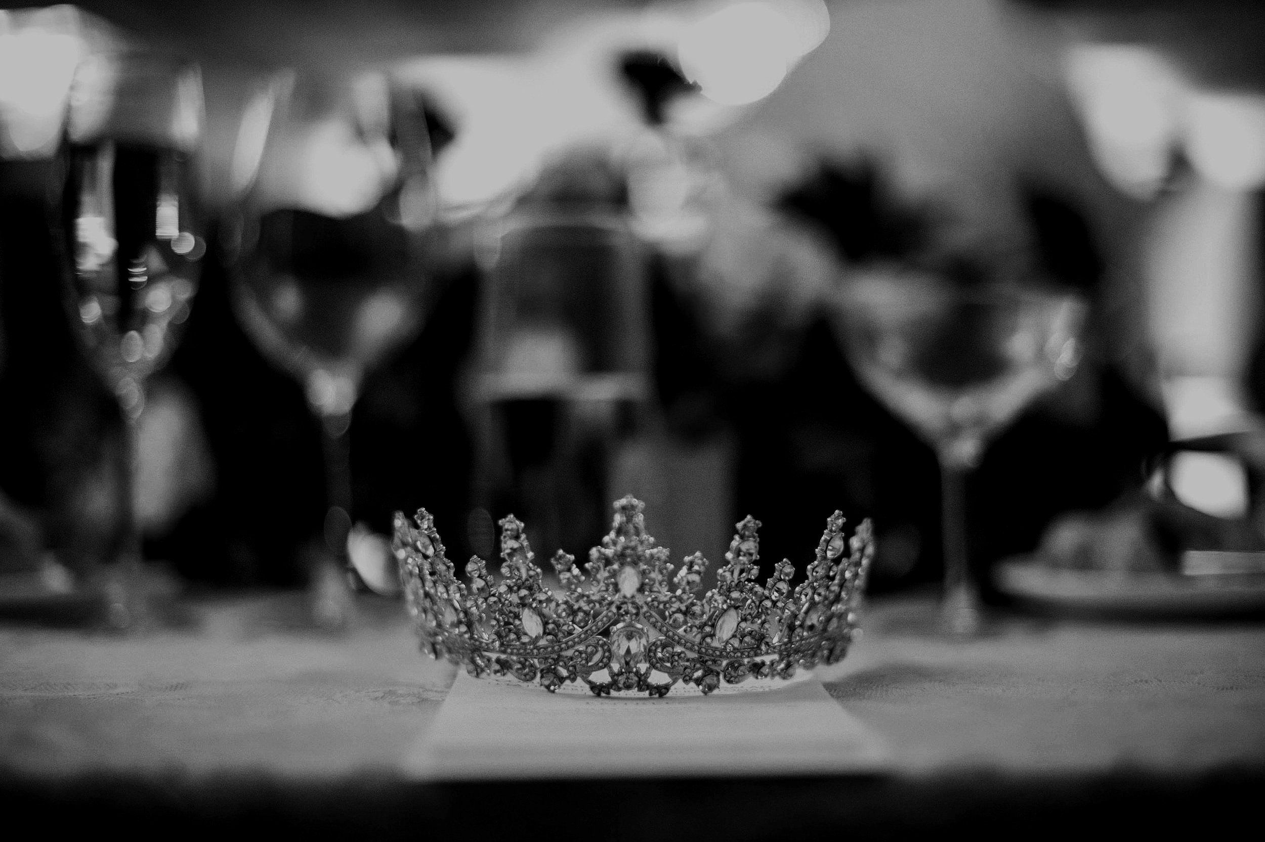 Black and white photo of a decorative tiara placed on a table in front of blurred glasses.